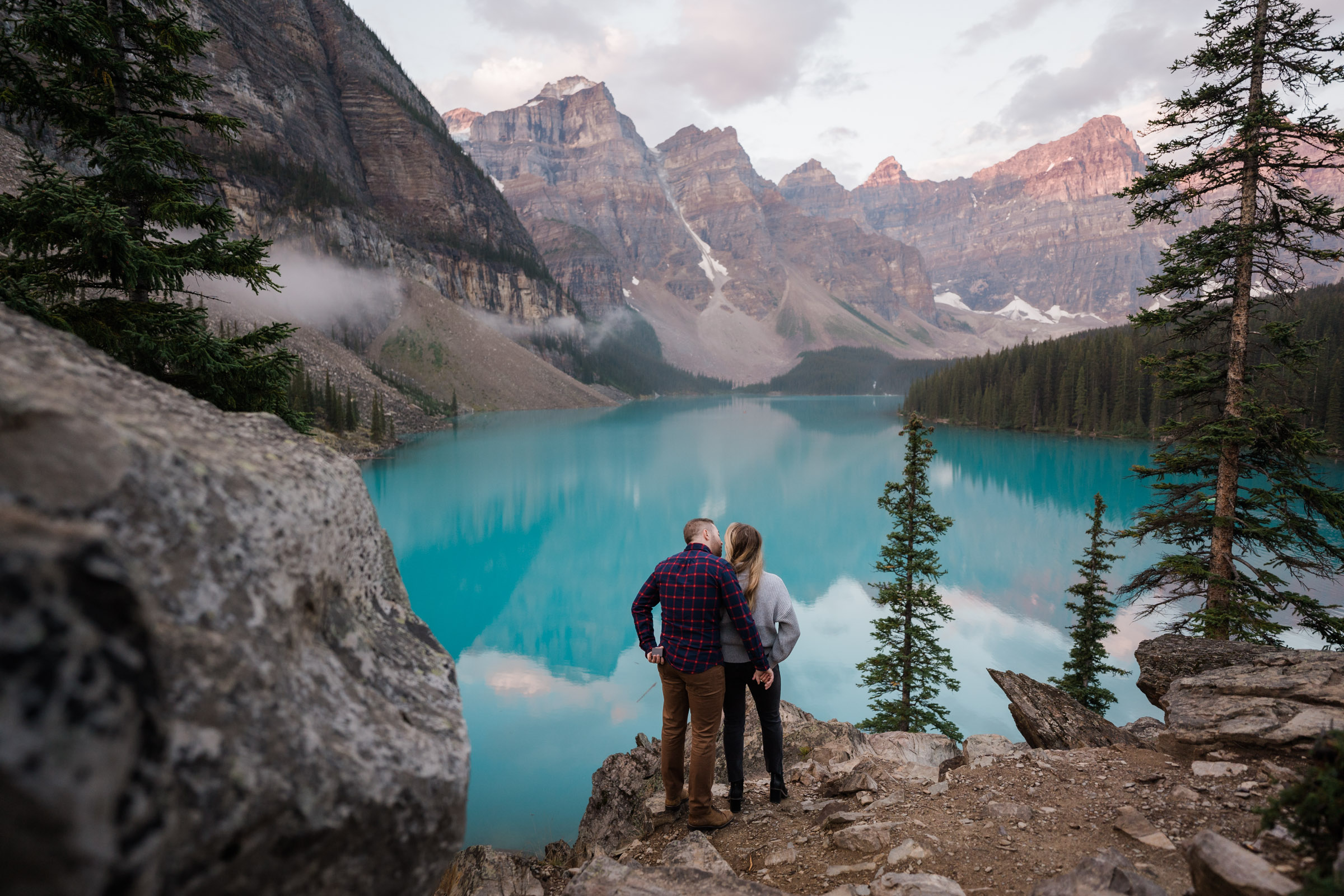 Couple kisses in front of Moraine Lake while he holds a hidden ring box behind his back