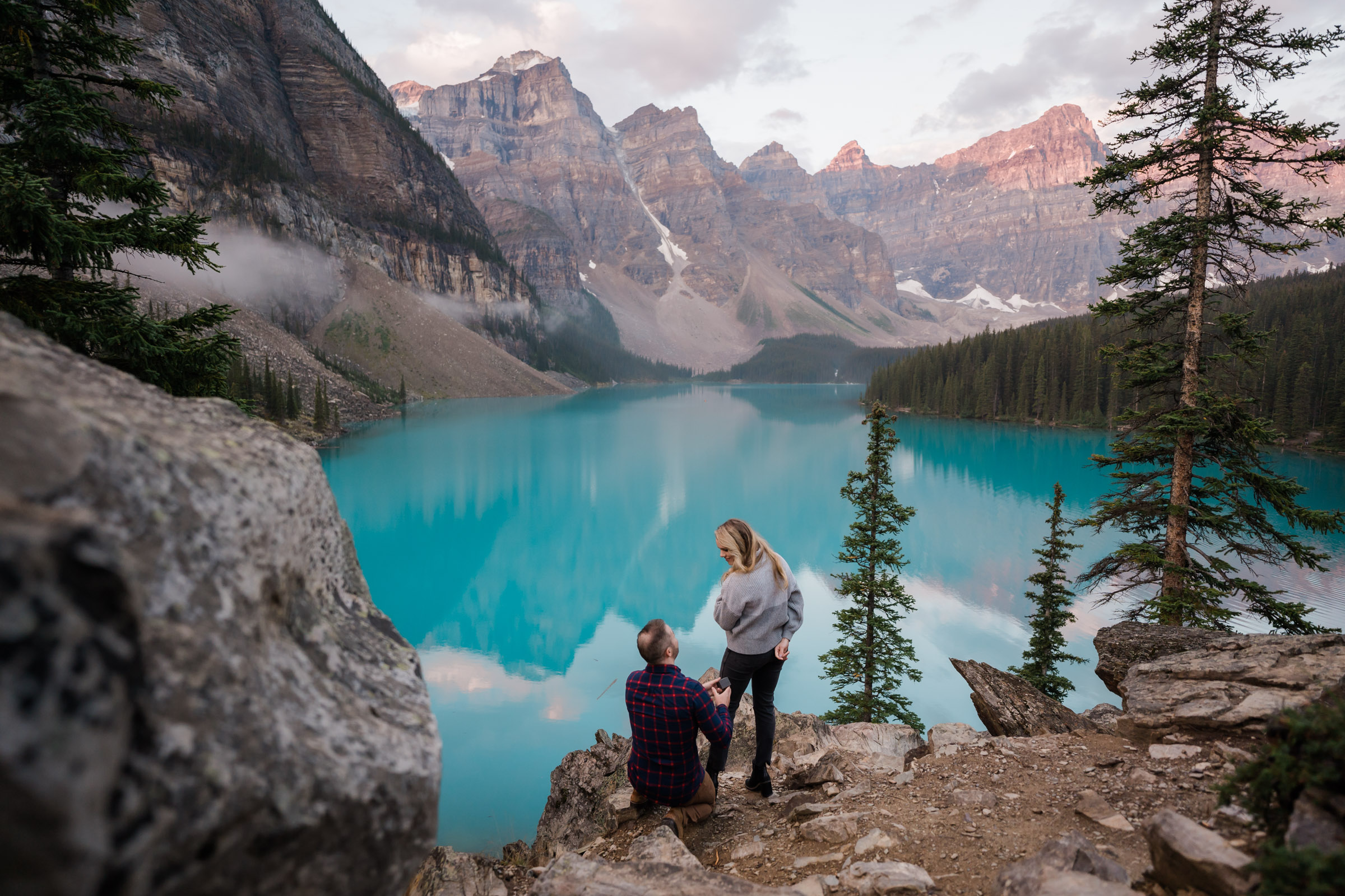 Man kneels on the ground while his girlfriend is turning around to see him proposing in front of sunrise at Moraine lake.