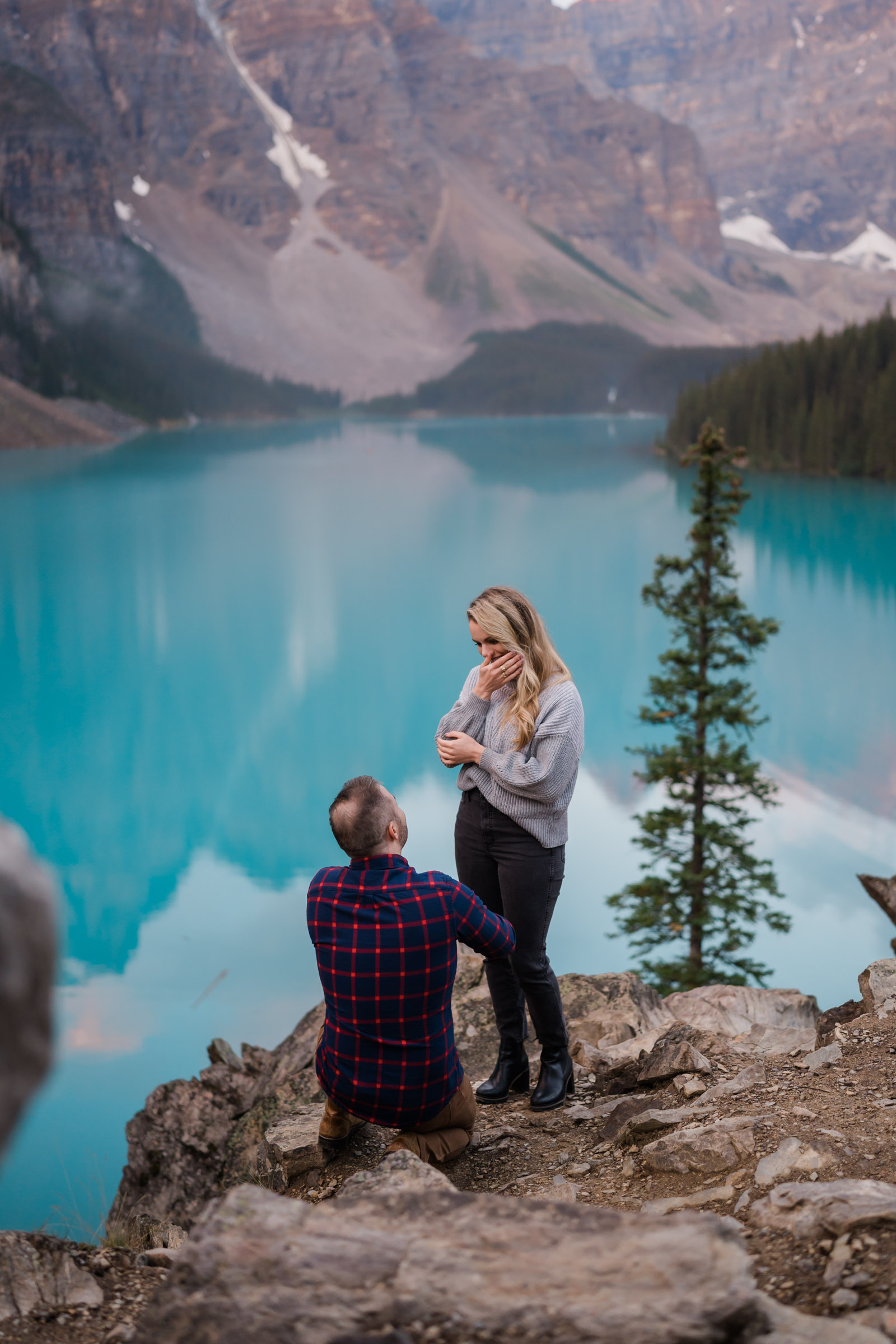 Woman standing in front of the turquoise blue of Moraine Lake as her boyfriend proposes. 