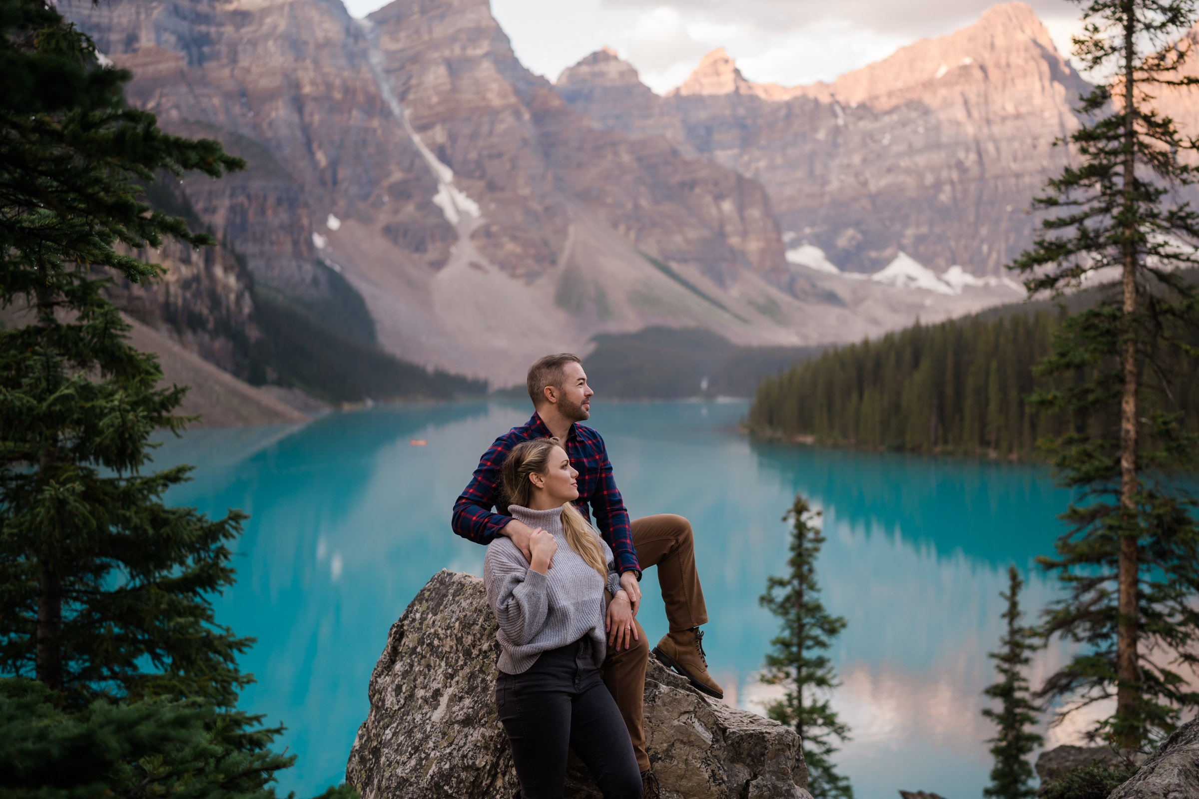 Man sits on rock as the sun comes up over Moraine Lake with new fiance standing infront of him as they look off into the distance.