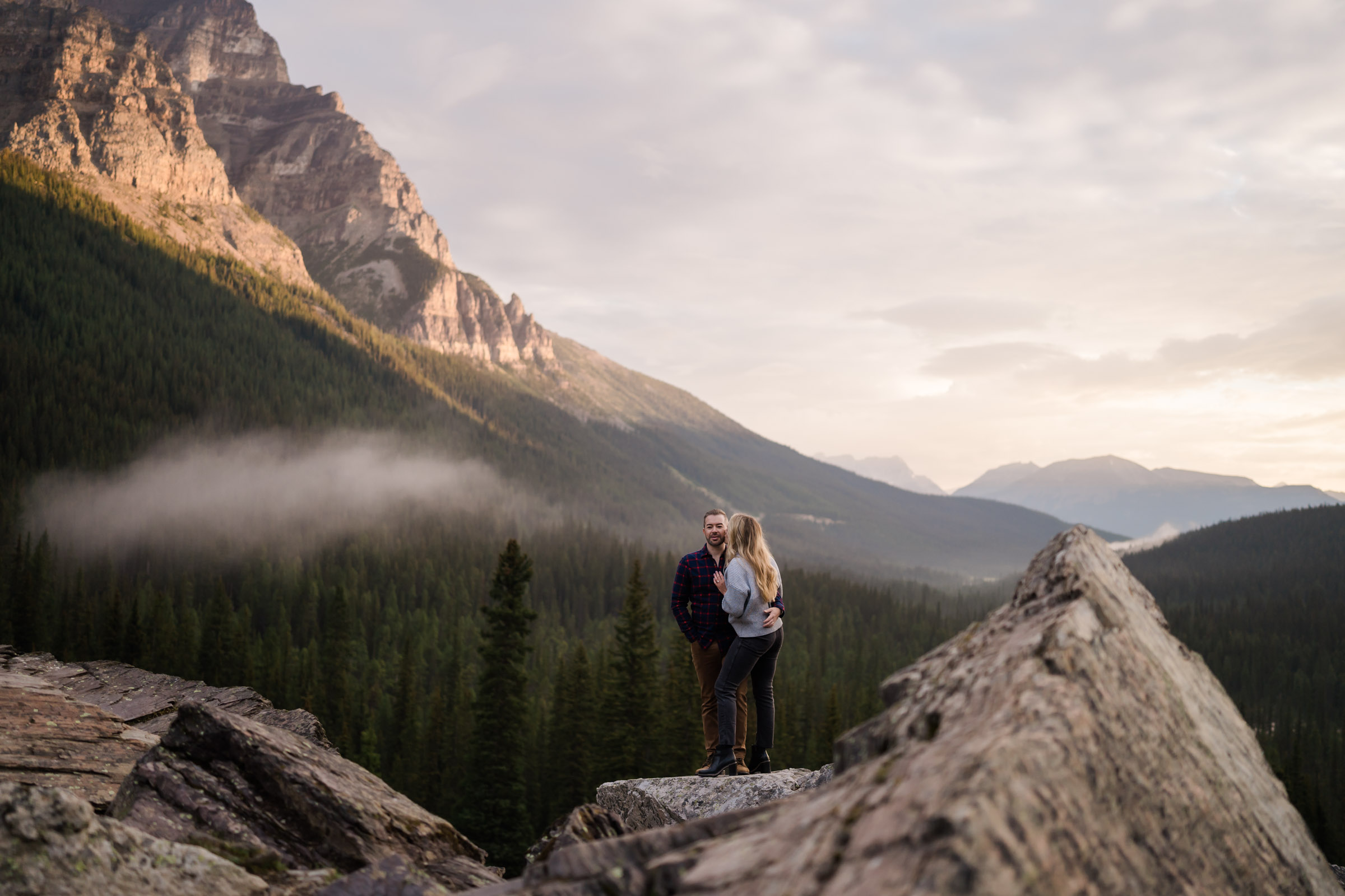 Fog floats over the valley as the sun lights up the mountains behind as a couple snuggles in Banff.