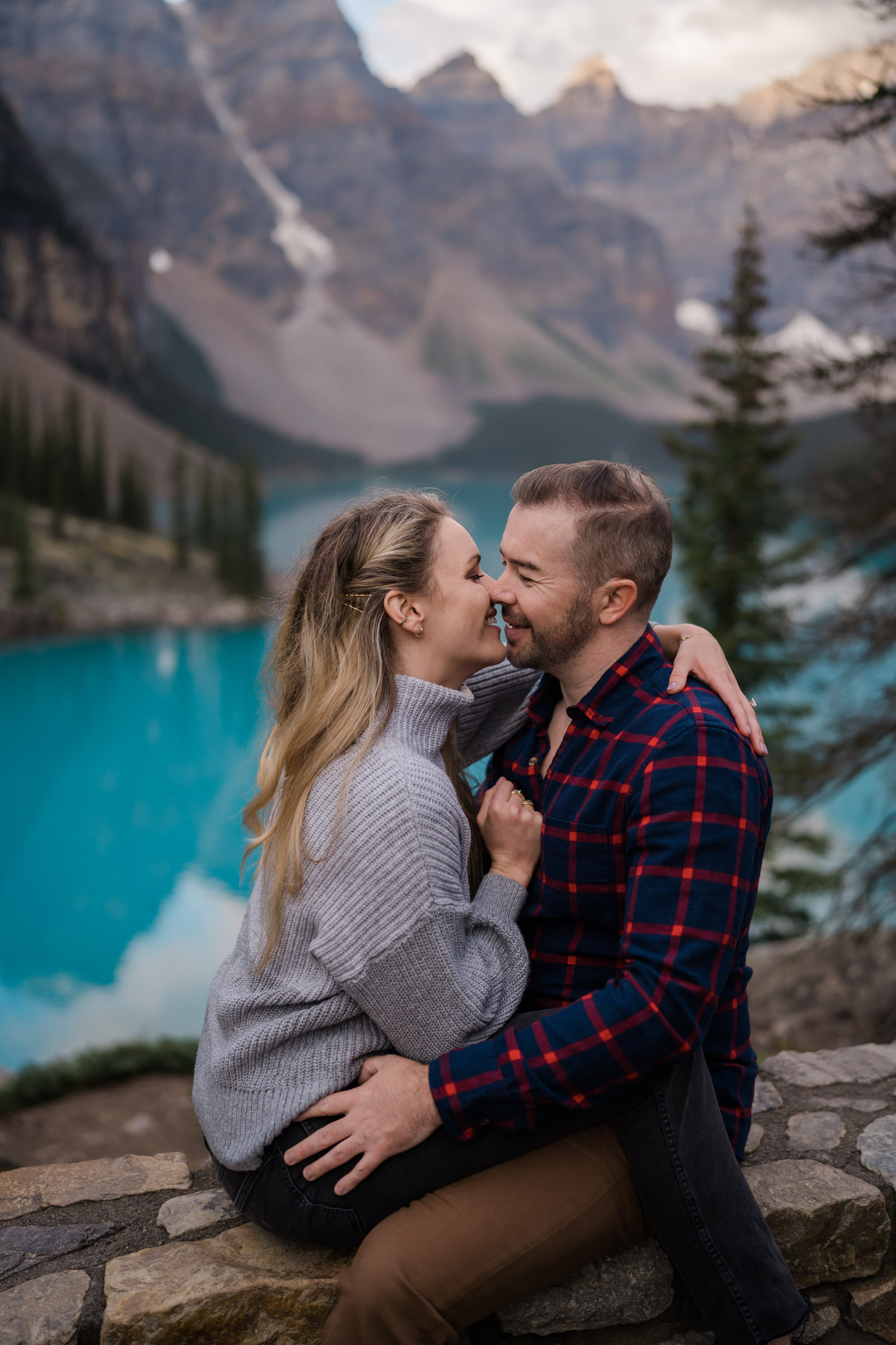 Couple sits facing each other and smiling almost kissing during Banff Engagement Photos. 
