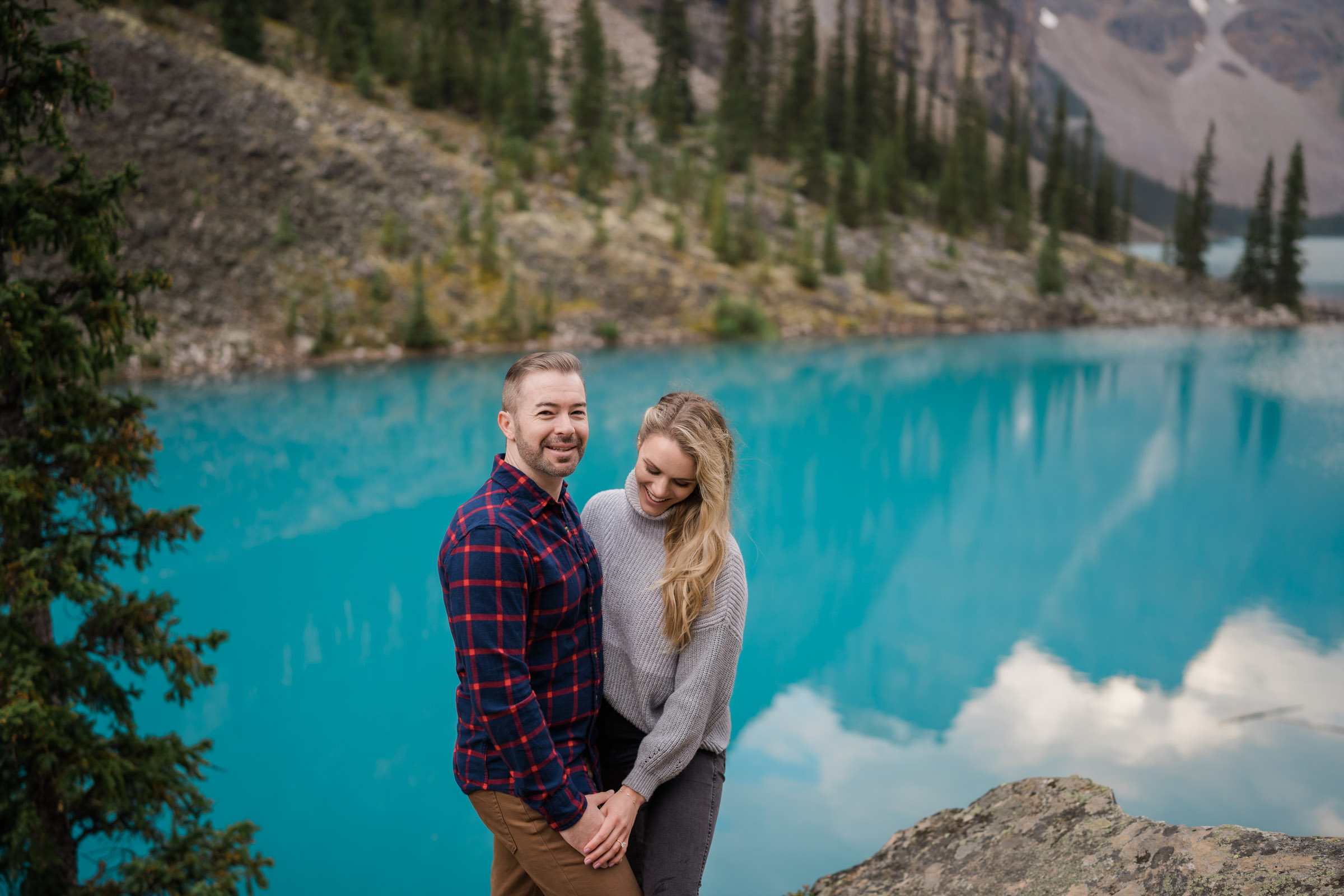Couple laughs as she looks down at her new ring and he smiles at the camera in front of the turquoise blue of Moraine Lake.