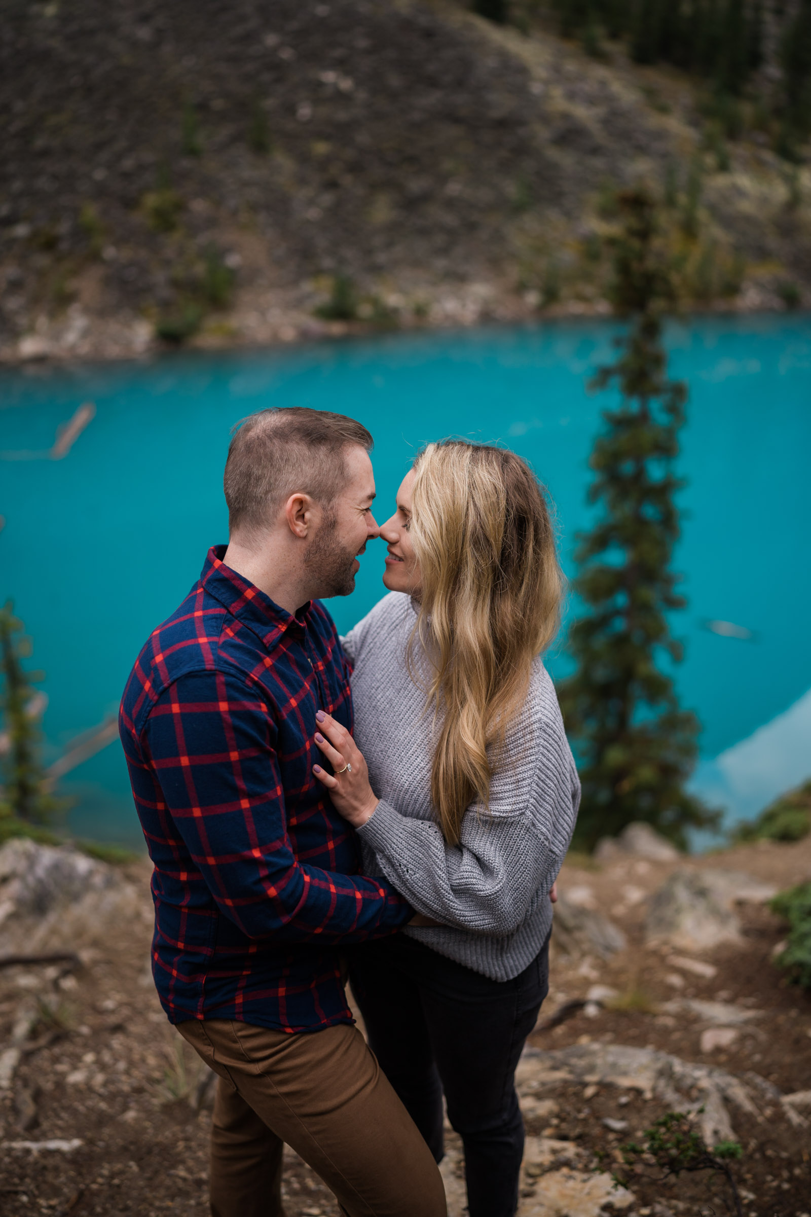 Close up of man and woman going in for a kiss with the bright blue water of Moraine Lake behind them.