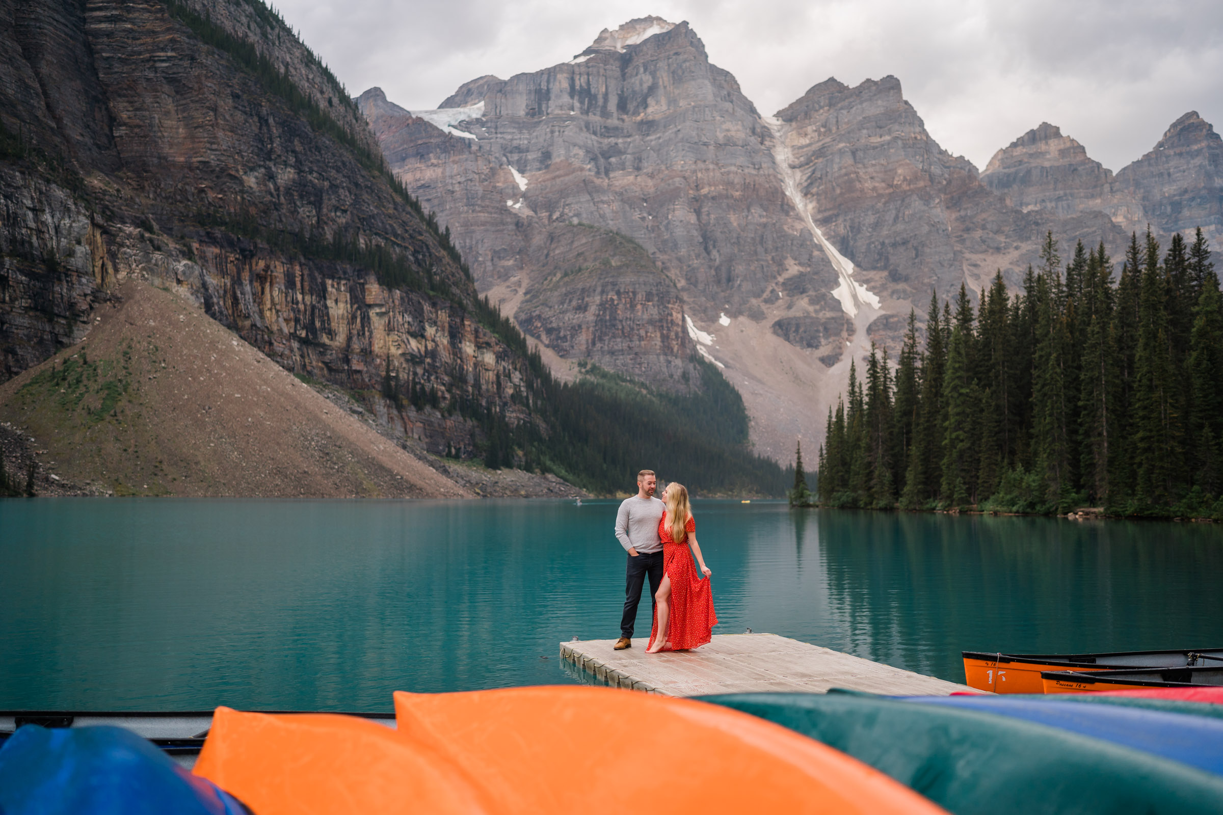 Couple stands on dock at Moraine Lake while she holds her red dress out to the side with the multi coloured canoes in the foreground. 