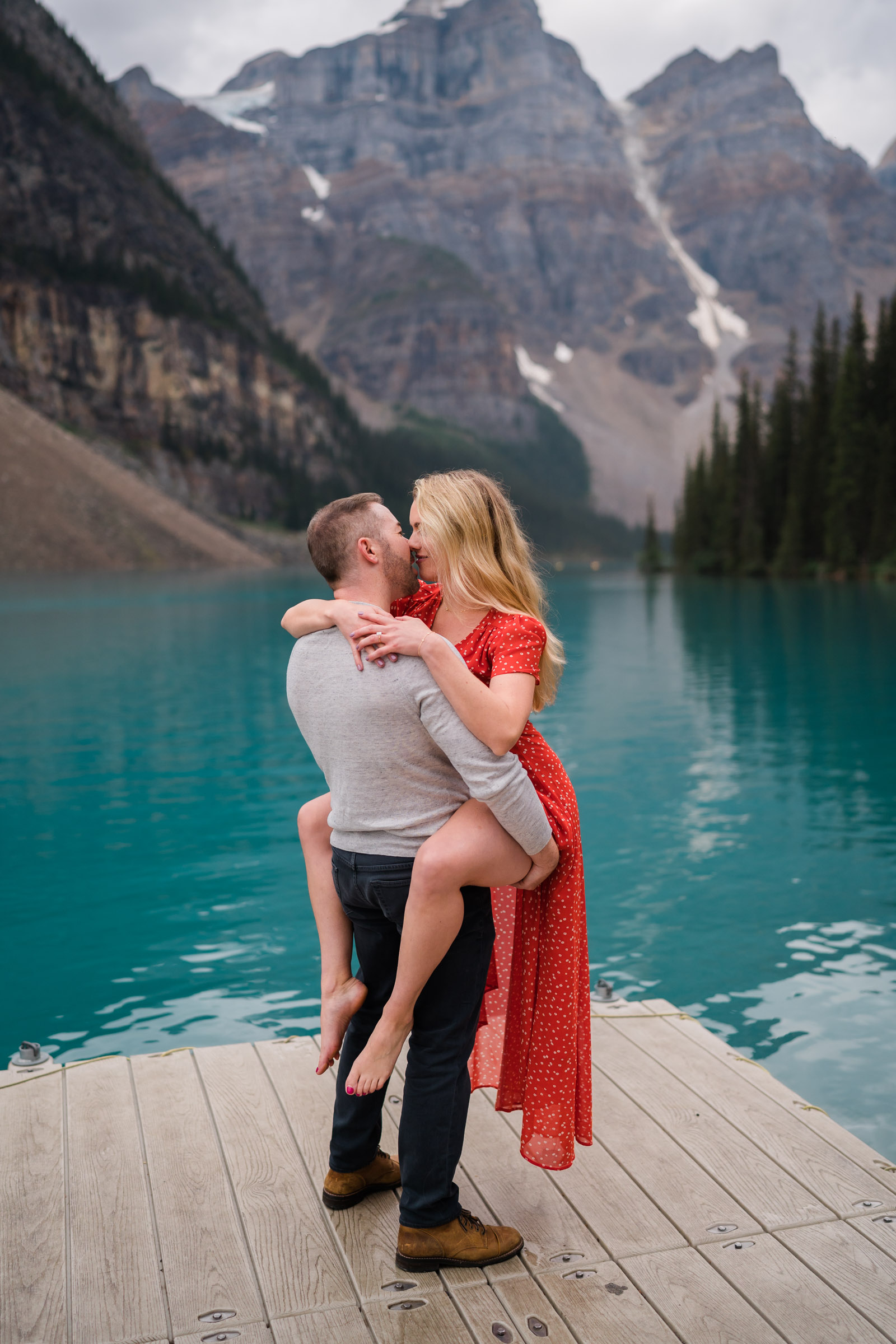 Man holds new fiance up as she wraps her legs around him while wearing a red dress on the docks at Moraine Lake. 