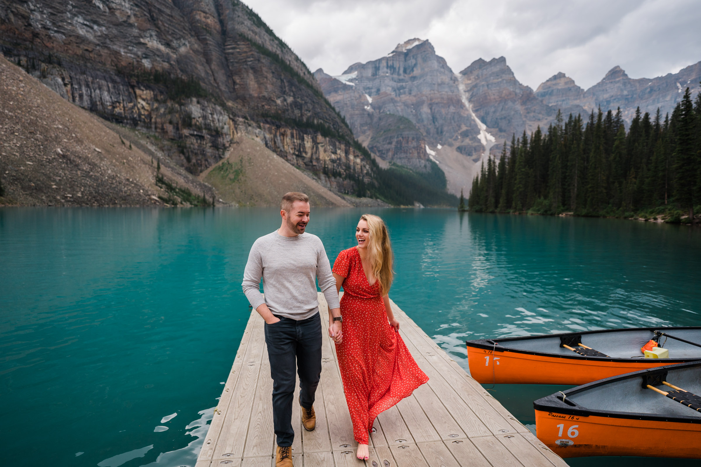 Man and woman holds hands while walking and laughing on the docks at Moraine Lake during their Banff Engagement Photos. 