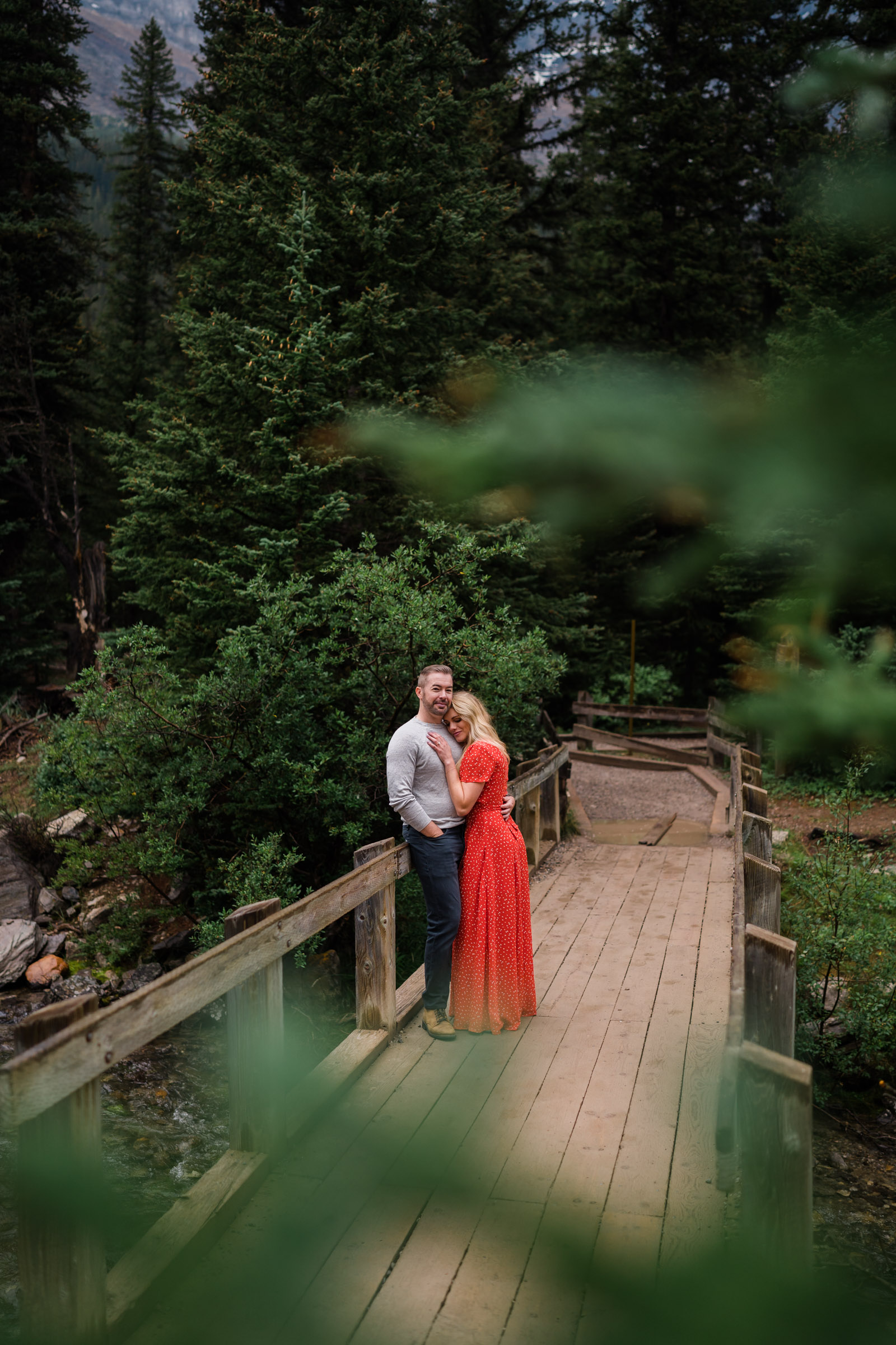Couple cuddles on wood bridge being framed by pine trees. 