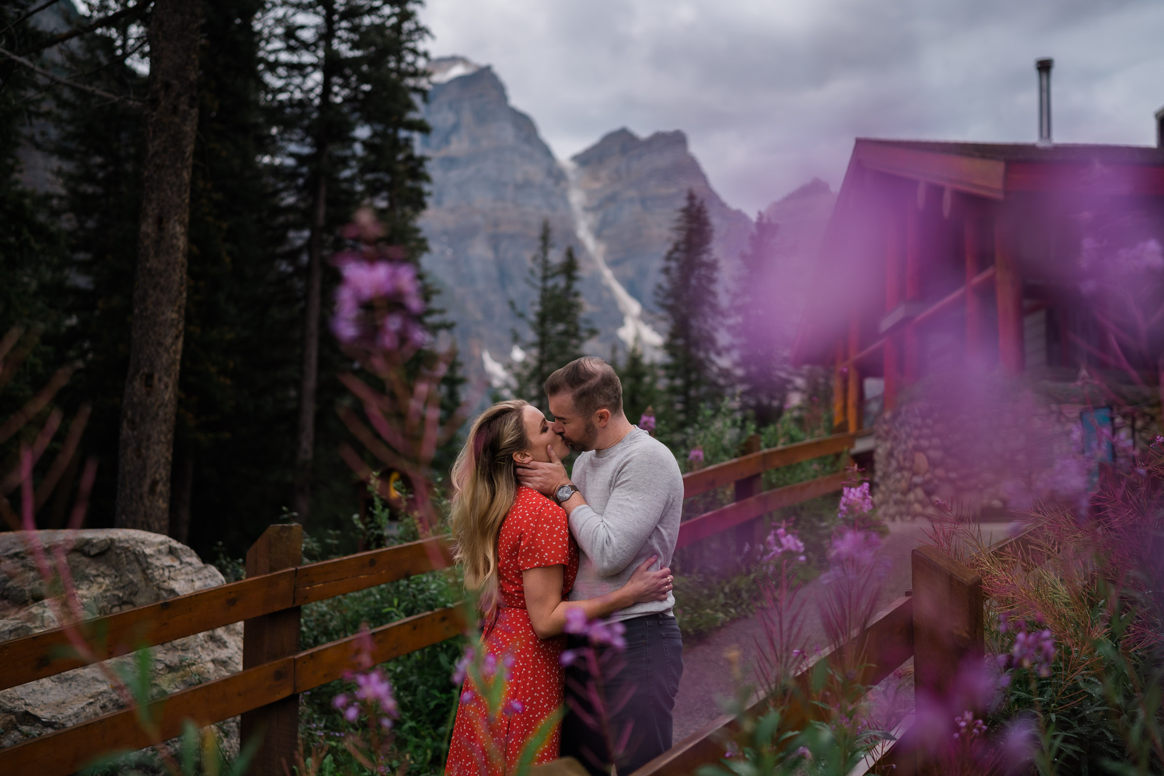 Couple kisses in front of Moraine Lake lodge in front of the purple blooms of the fireweed during their Banff Engagement Photos. 
