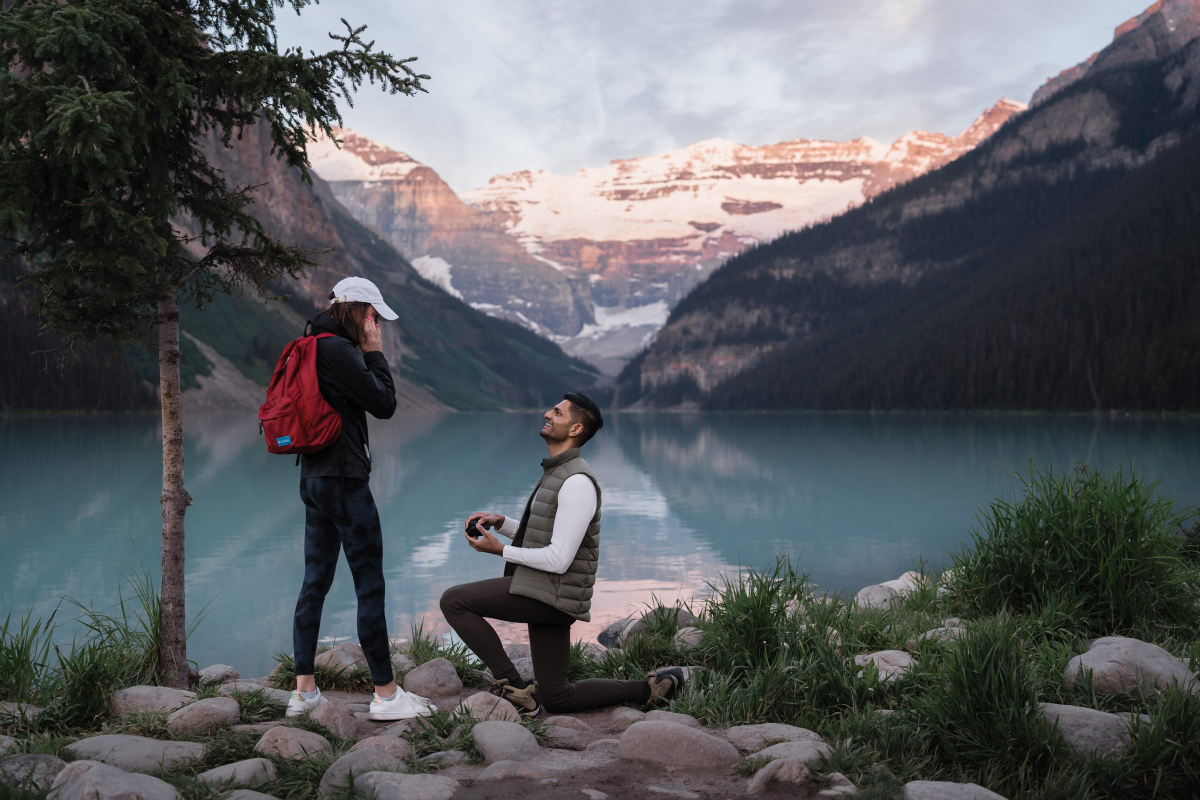 Man is down on one knee infront of surprised girlfriend as the sun lights up the glacier behind Lake Louise.