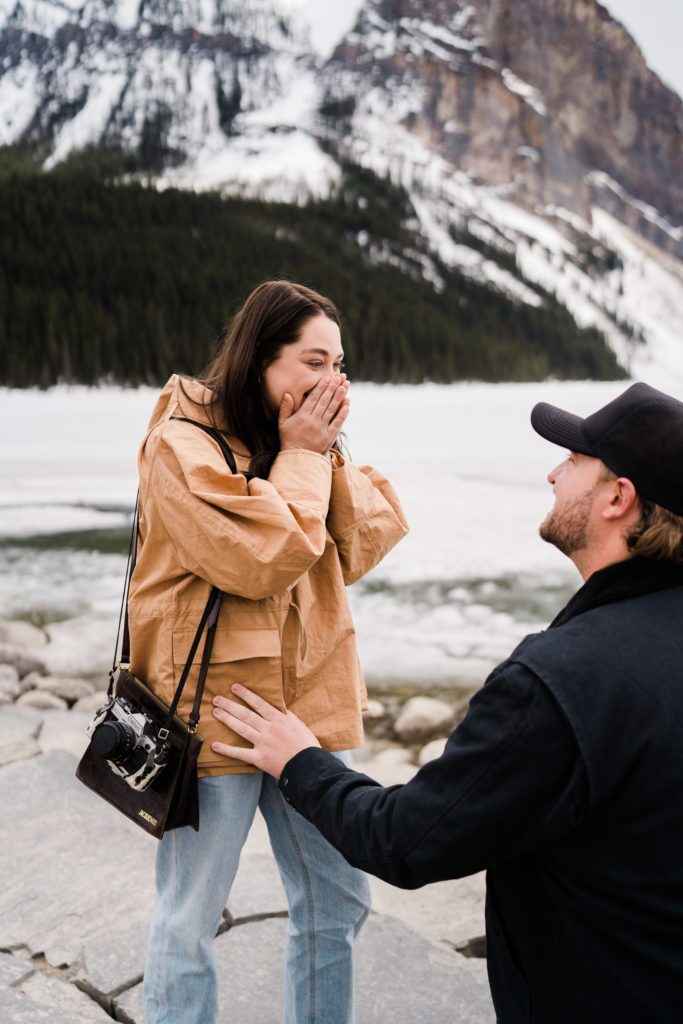Man is on one knee in front of girlfriend who is covering her face with surprise after a proposal. 