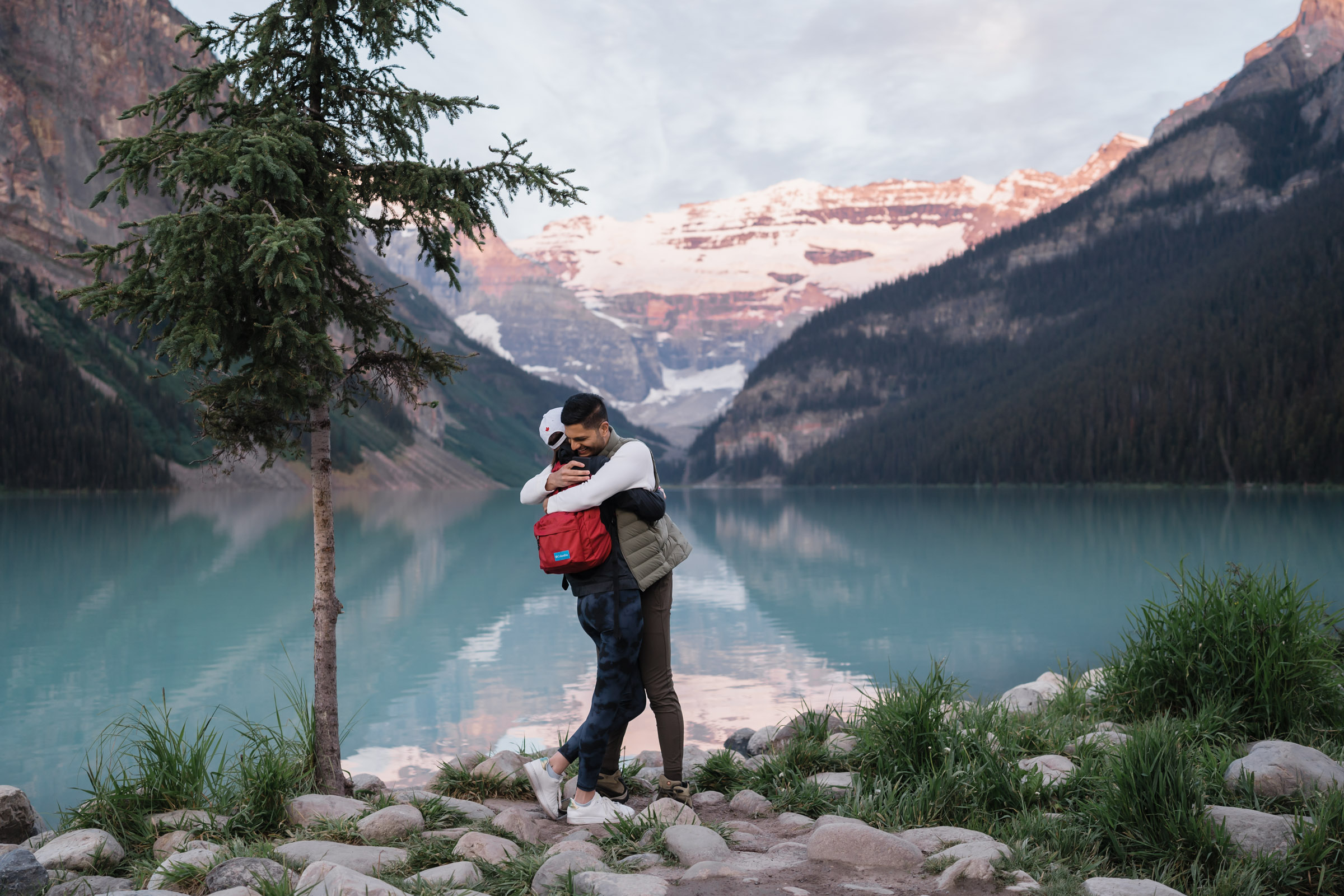 Man and women embrace after a proposal in front of Lake Louise at sunrise.