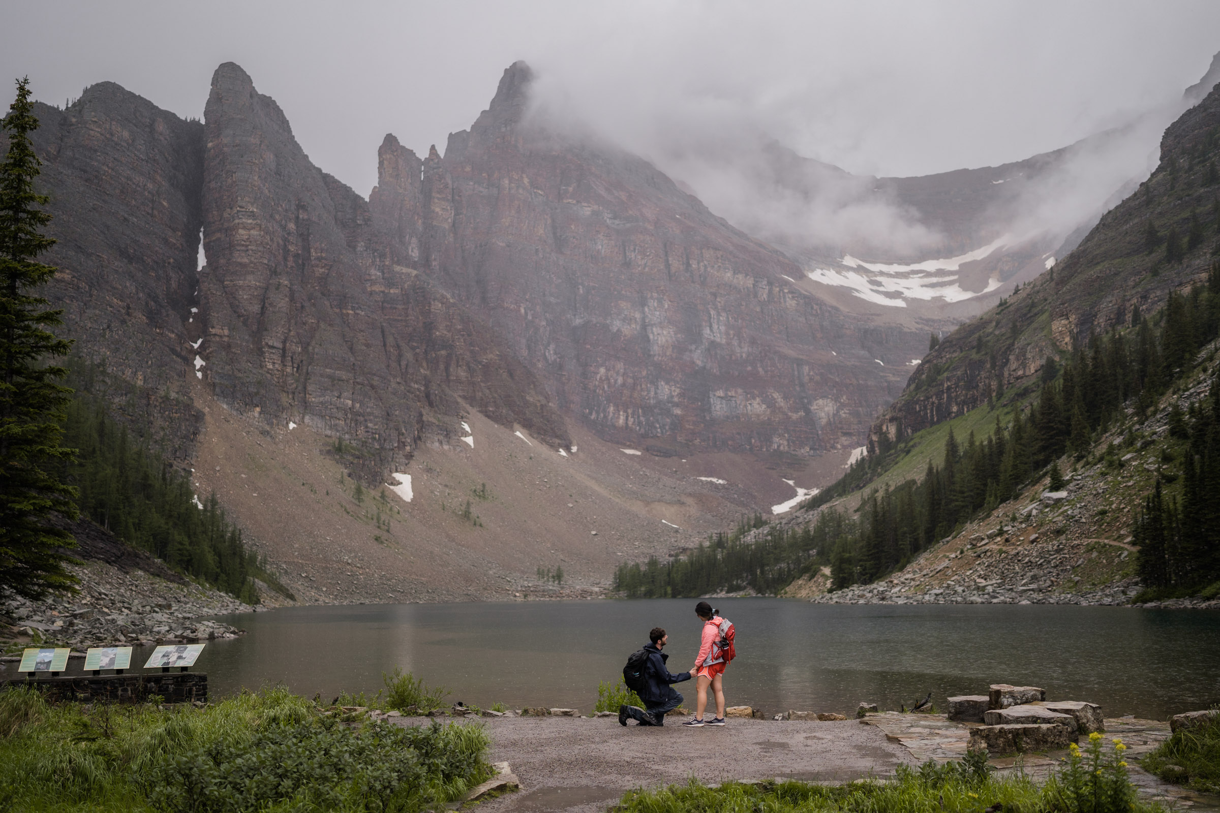 Man kneels down infront of girlfriend wearing pink jacket infront of Lake Agnes to propose in Banff National Park