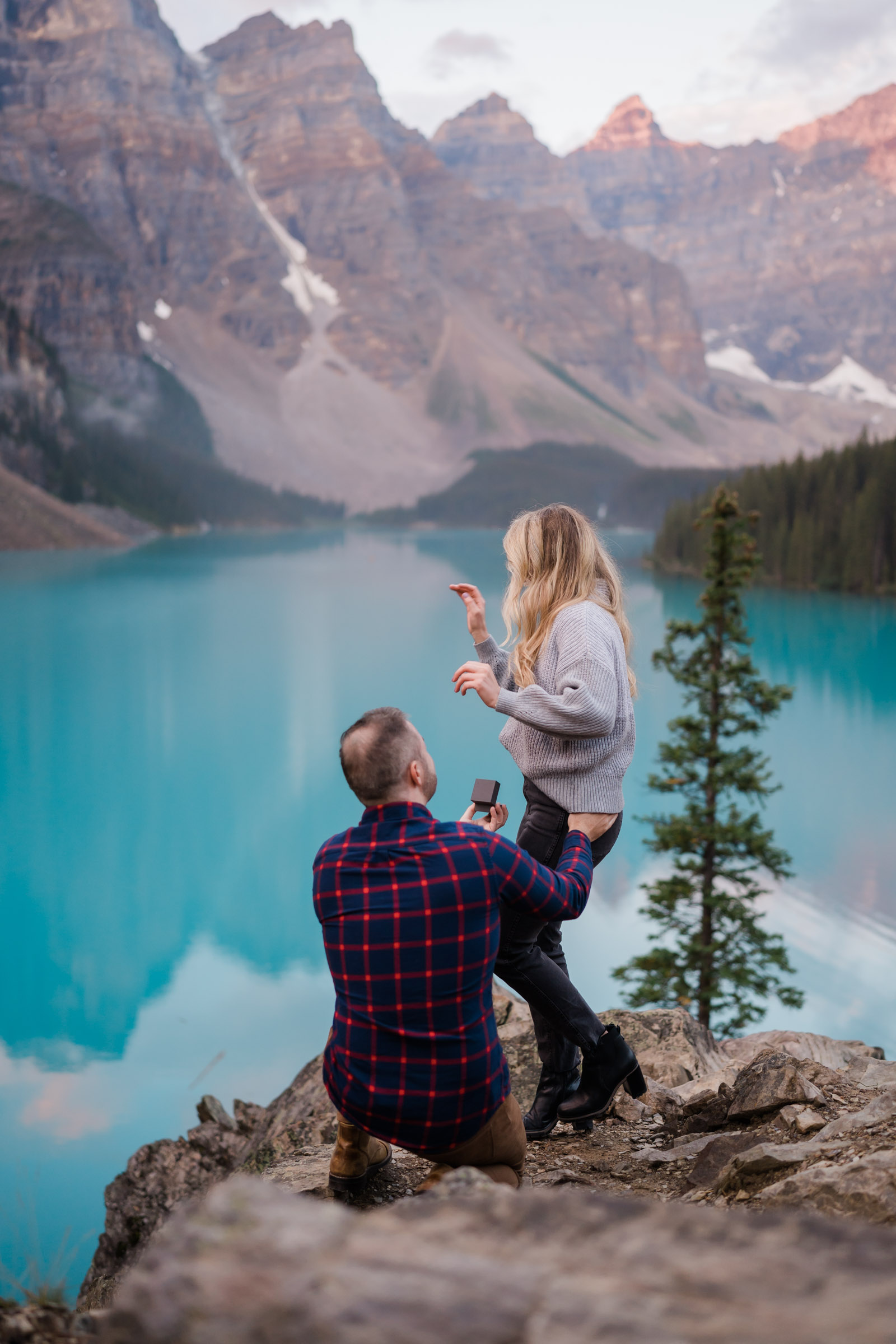 Man in plaid shirt is kneeling infront of his girlfriend showing her the ring as he is proposing and she is turning away in surprise