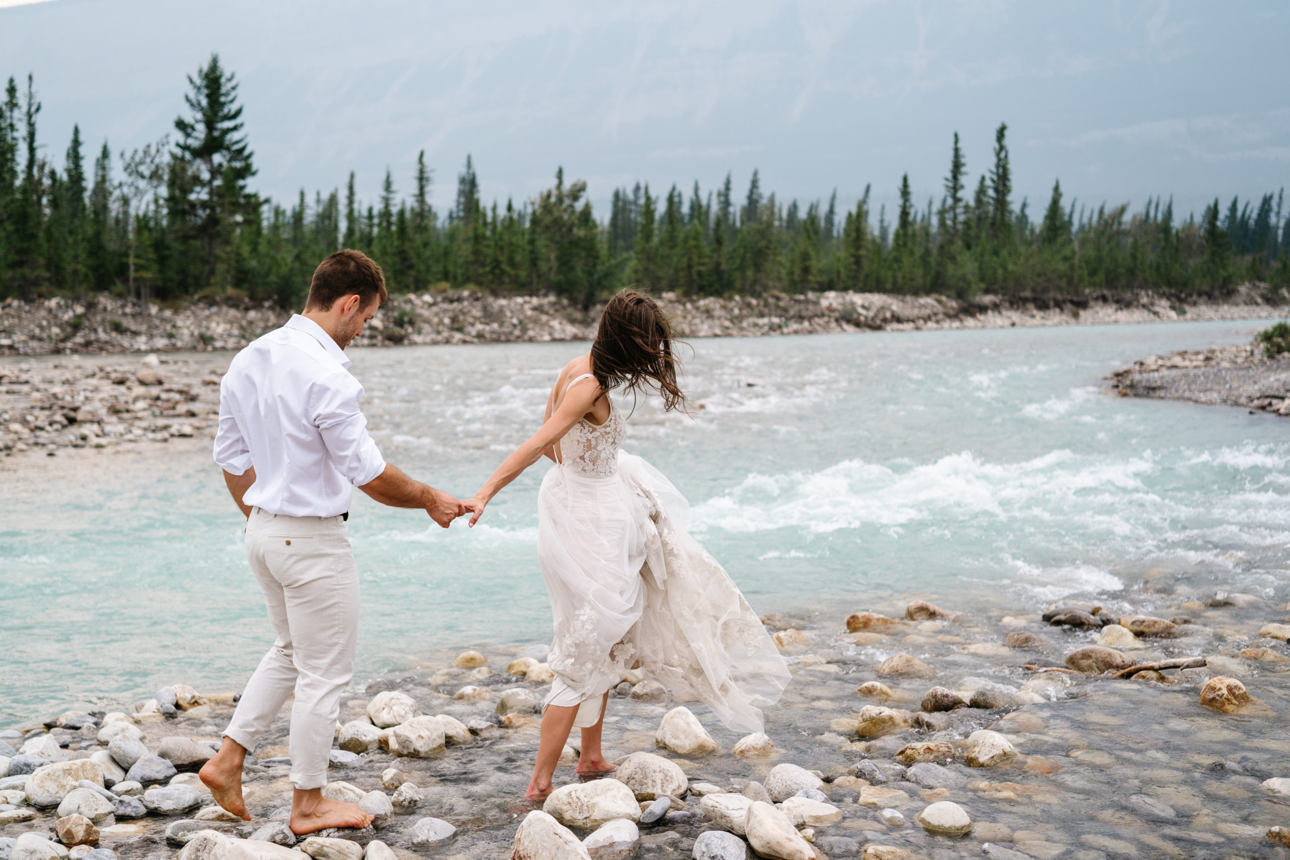 barefoot bride leads groom over rocks along the snaring river in jasper national park during their alberta elopement 