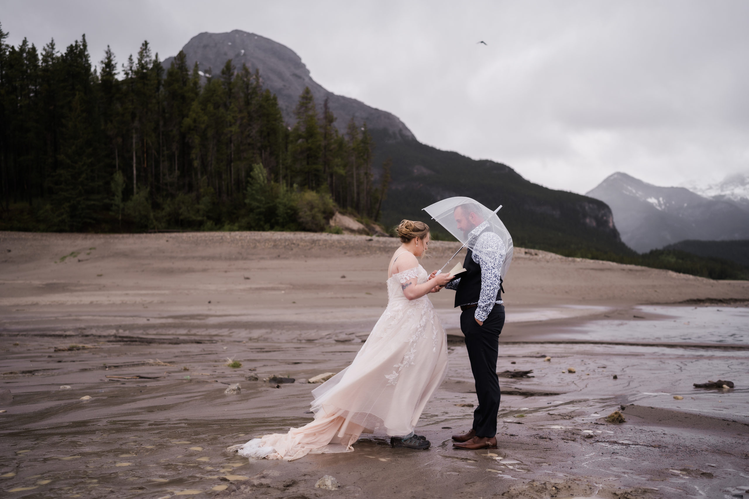 bride and groom stand facing each other reading vows on muddy beach with the mountains in the background while it rains during their kananaskis elopement at barrier lake