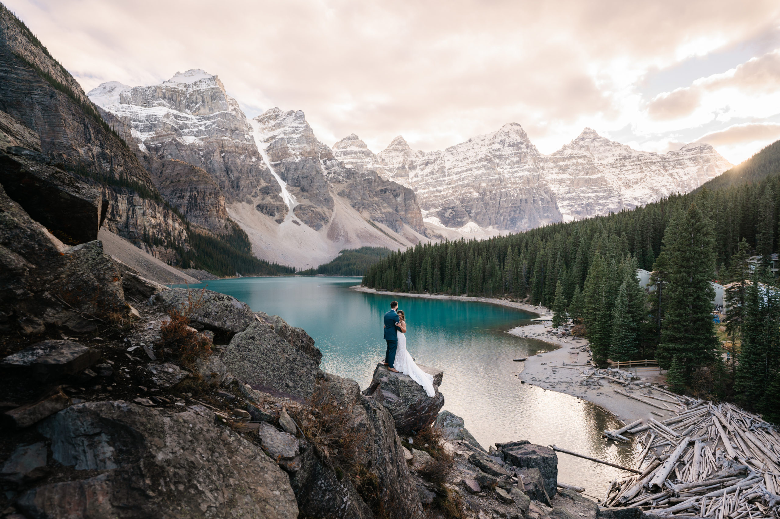 the sun sets over the valley of the ten peaks at moraine lake while a bride and groom stand together holding each other on a rock with the lake in the background 