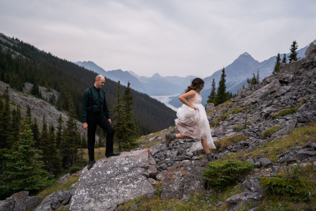bride in wedding dress and hiking boots jumps off rock that groom is standing on with mountain and lake in the background during a banff national park elopement