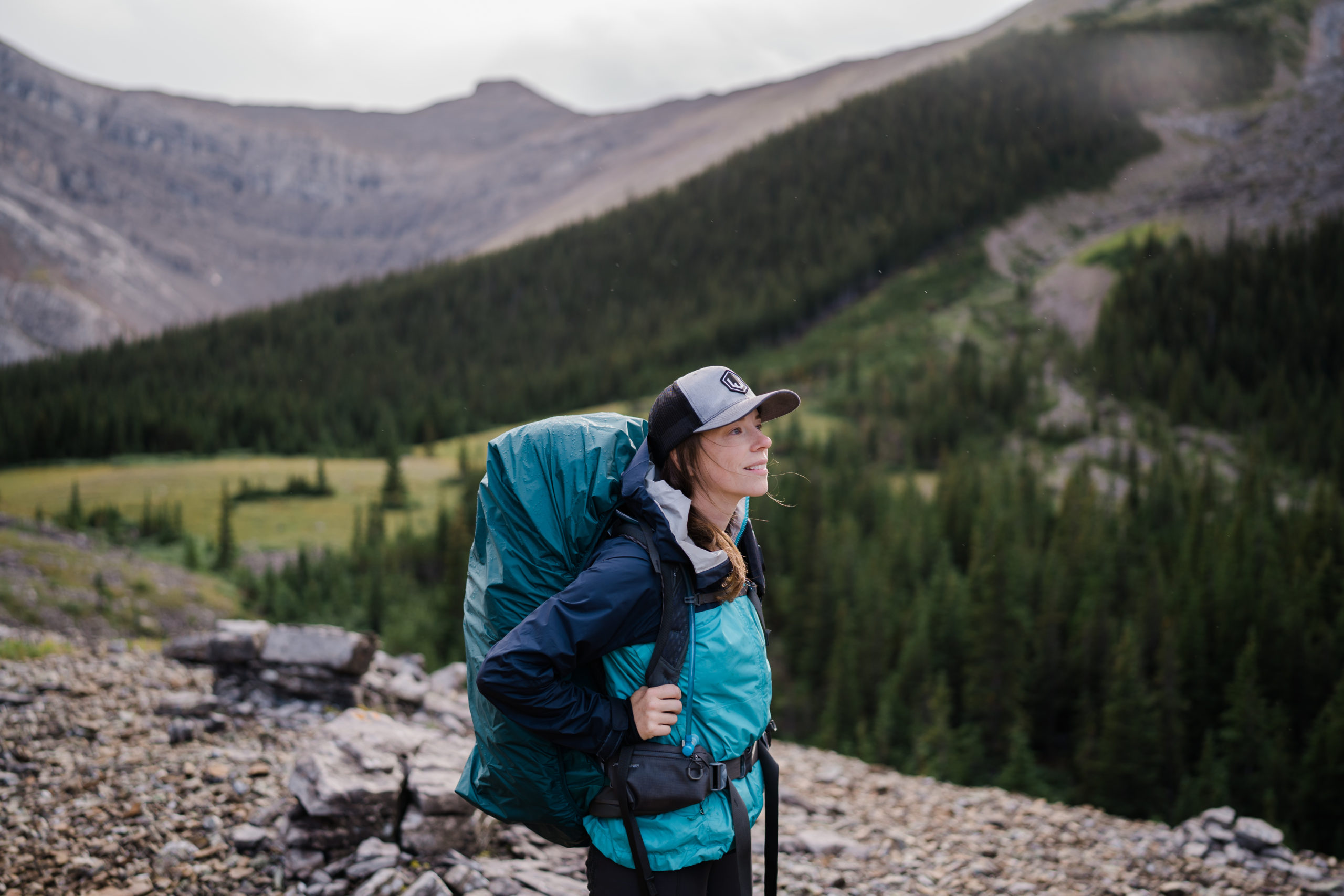 alberta elopement photographer wearing a grey hat and a blue jacket looks off into the distance as the sun sets on the mountain behind her while on a backpacking trip