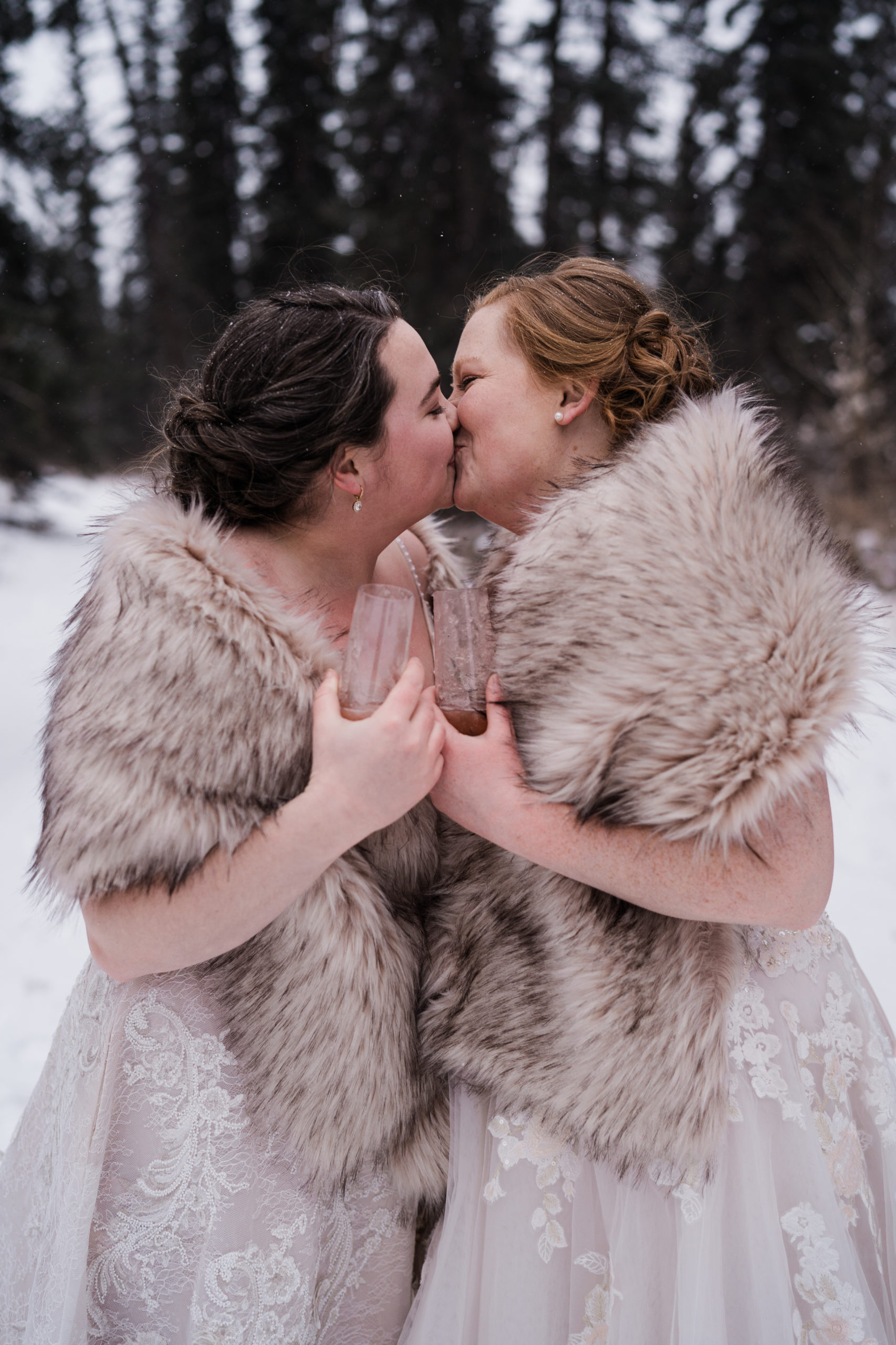 two brides wearing fur shawls kiss during their winter alberta elopement