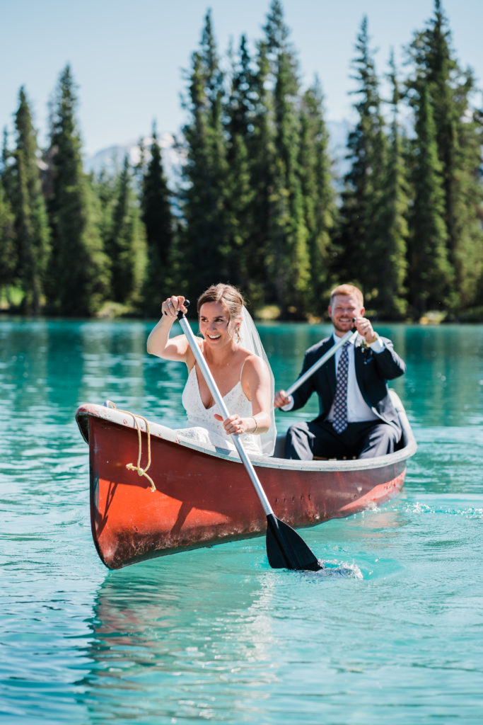 bride sits in the front of a red canoe while padding through blue water in jasper national park on their wedding day with the groom smiling in the back