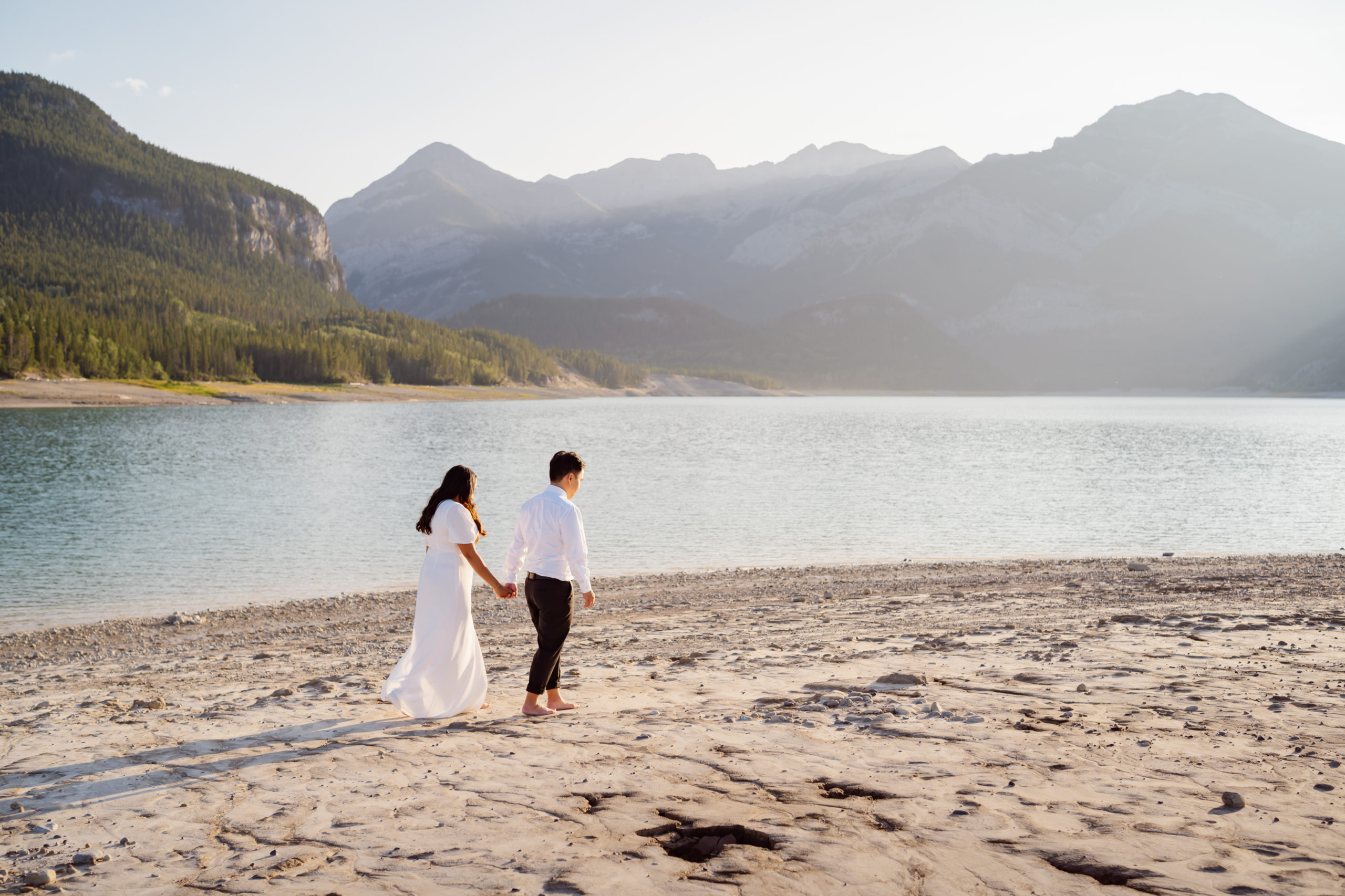 bride and groom walk barefoot down a beach at an alberta mountain like during the sunset after their elopement