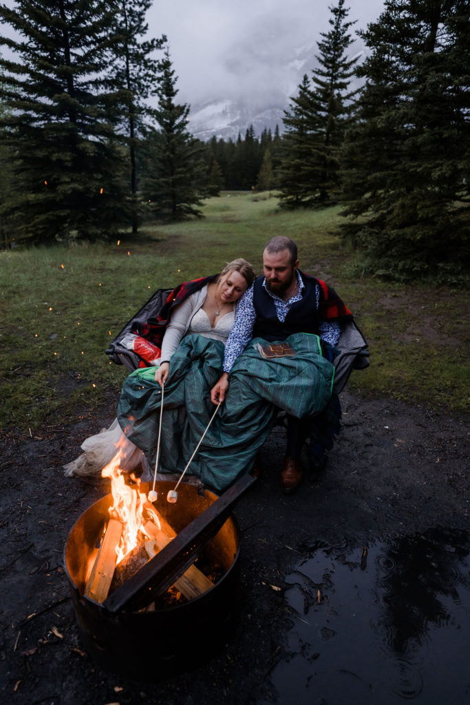 couple sits on chair wrapped up in blankets while roasting marshmallows in front of flaming fire on their wedding
