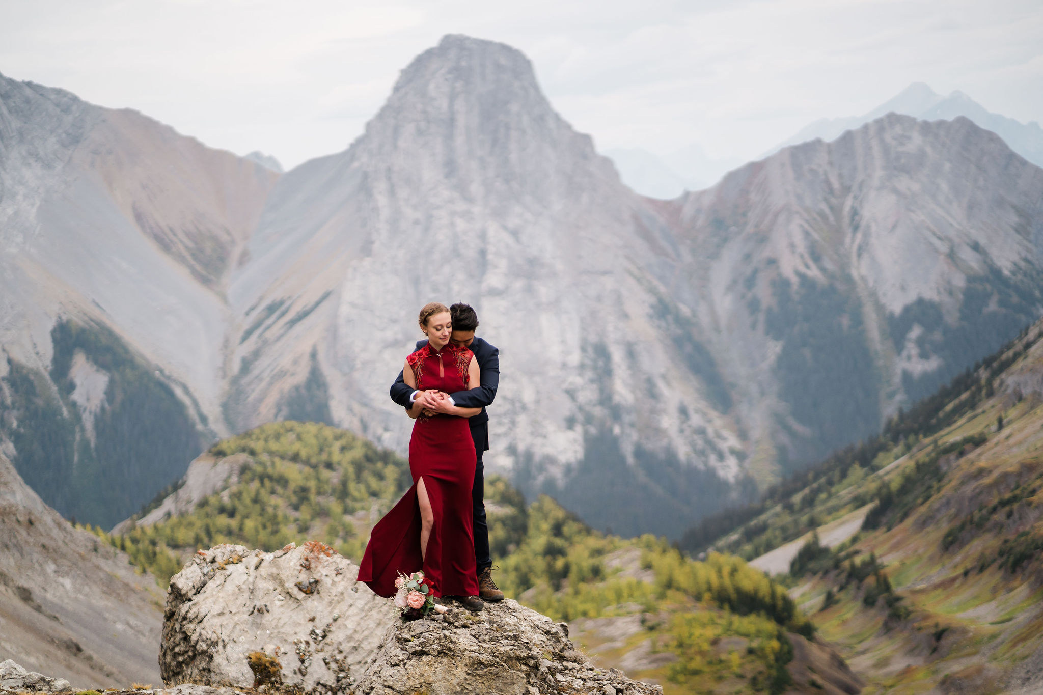 bride in red dress and groom cuddle on a rock with mountains and golden larches behind them during kananaskis hiking elopement
