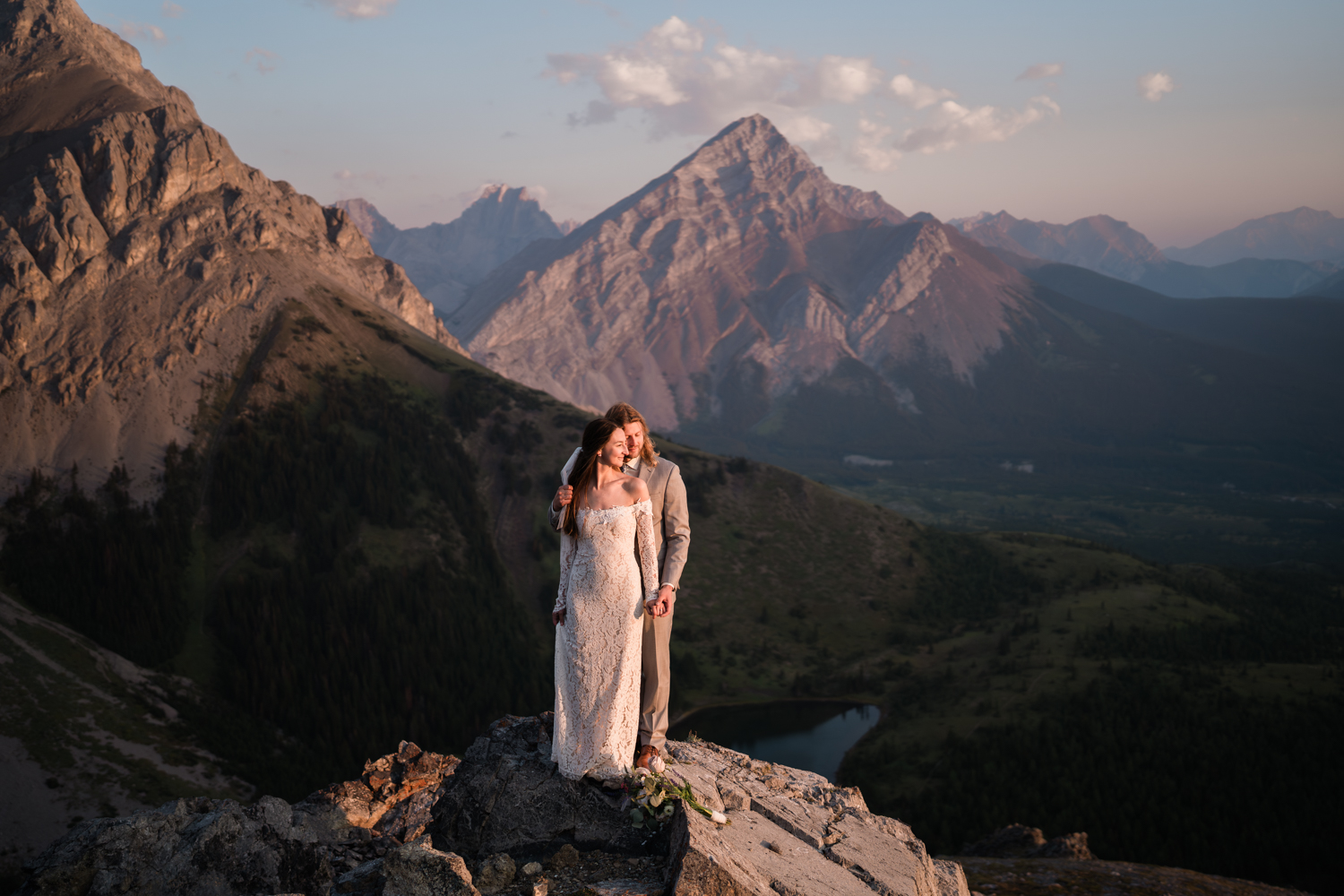 bride and groom stand on a rock overlooking a mountain lake as the sun comes up and casts warm light onto them