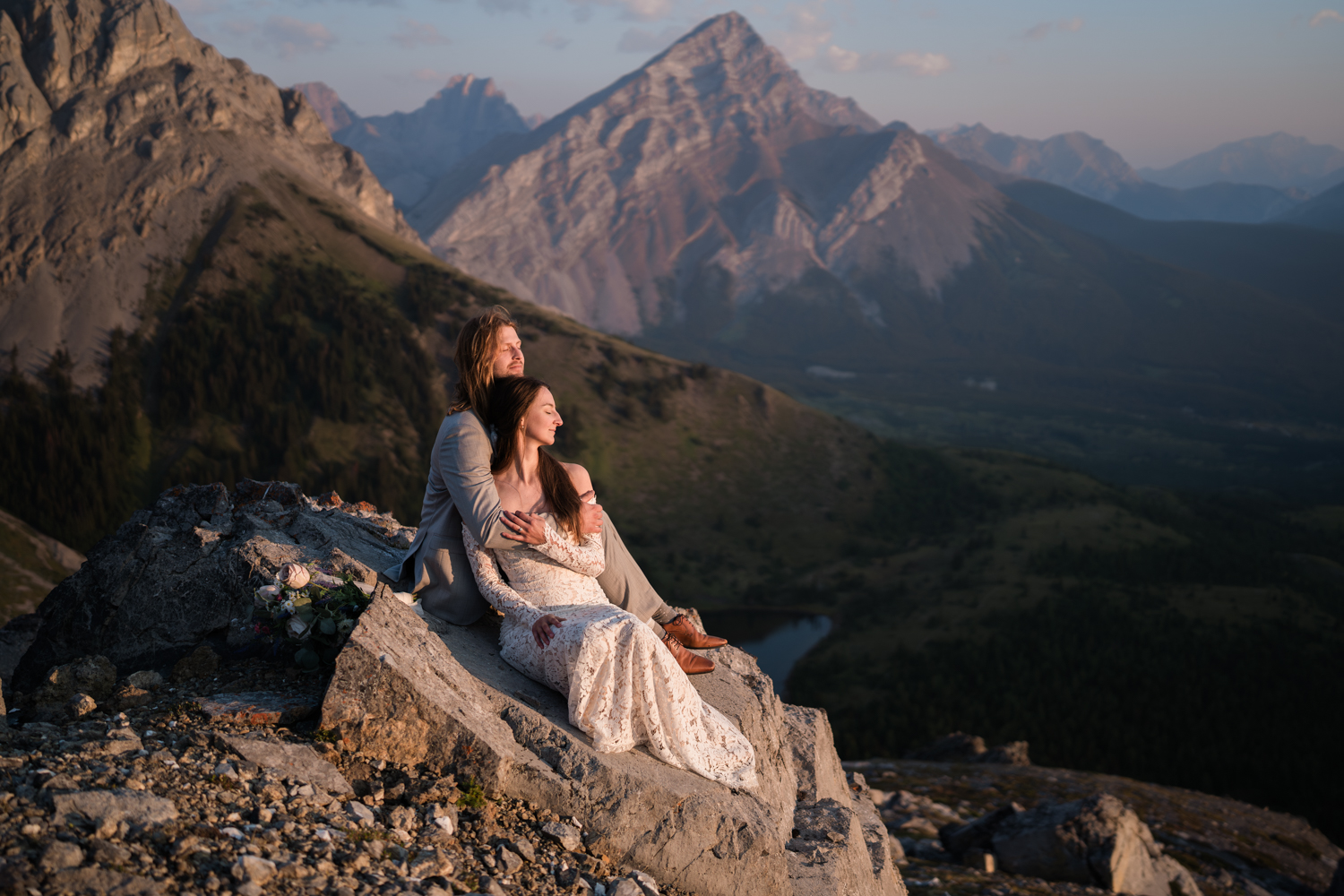 couple sits on a rock together eyes closed cuddling as the sun comes up and shines on them with mountains in the background during a kananaskis elopement.