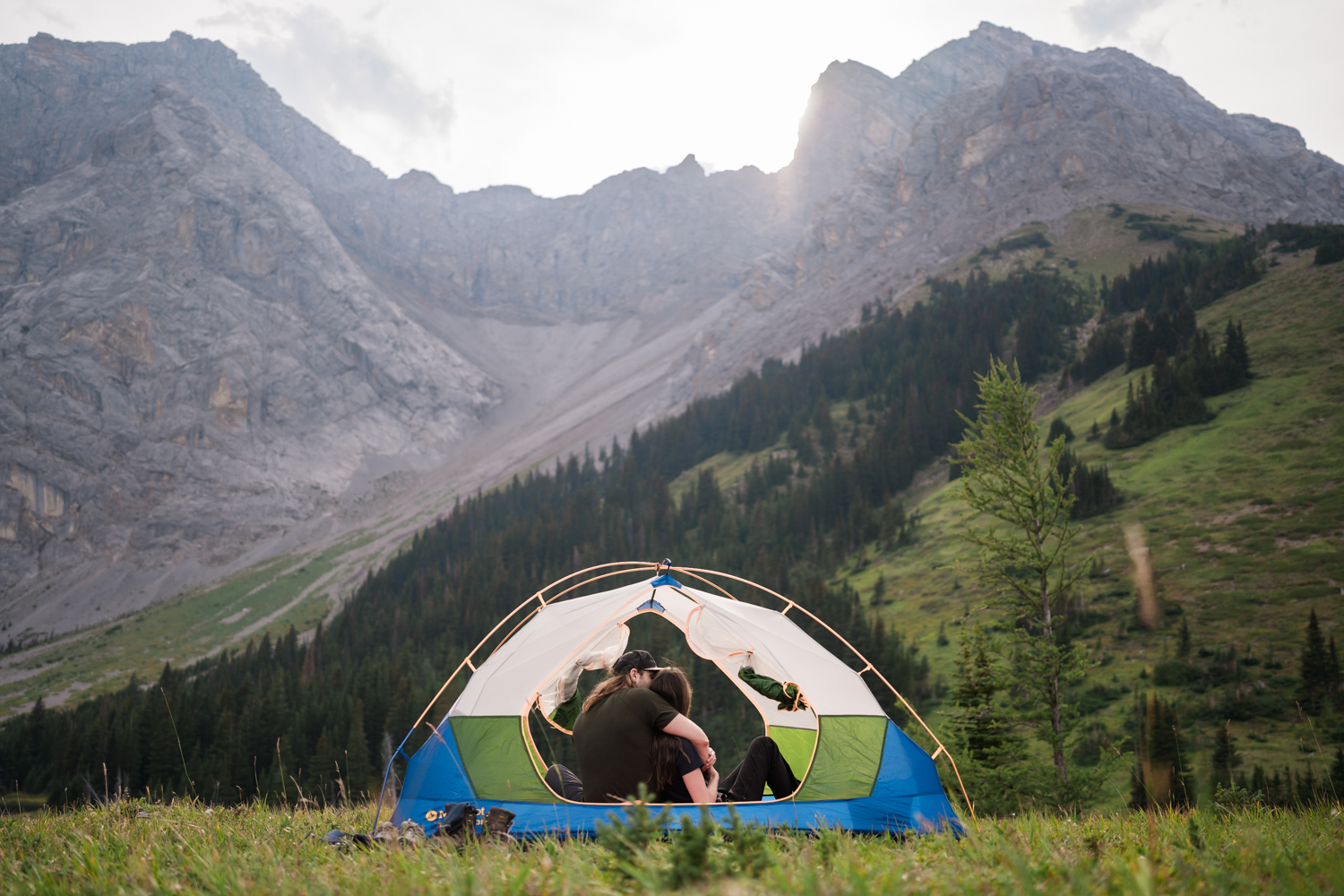 couple cuddles inside of green white and blue tent while the sun goes down behind a mountain in front of them during their camping elopement