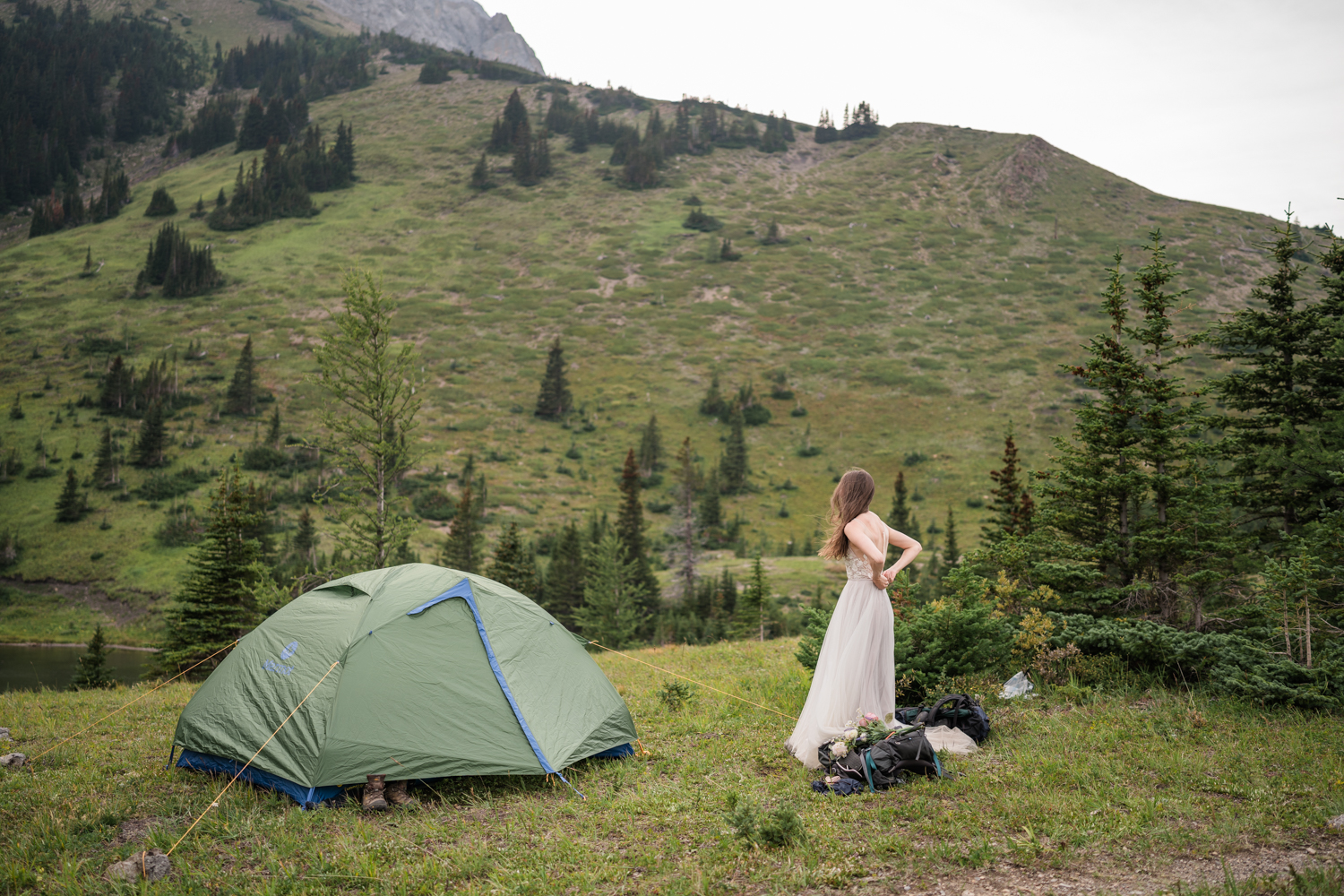 bride does up her wedding dress while standing beside a green tent in the mountains before a camping elopement