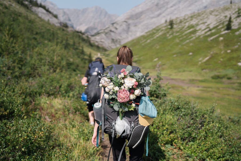 silk flower bouquet in a backpack while a bride walks along a green trail in the mountains