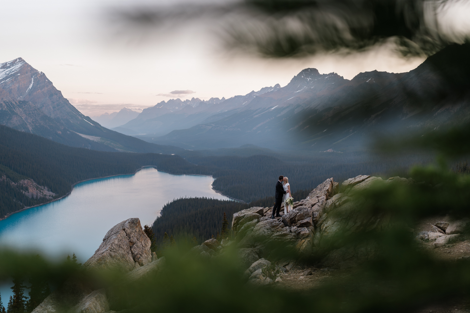 A look through the trees at a bride and groom embracing at Peyto Lake in Banff National Park