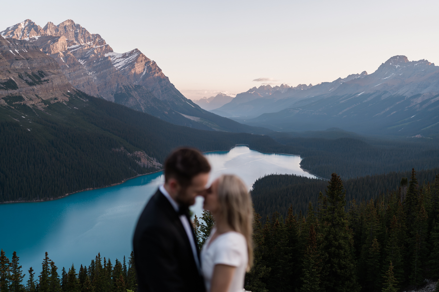 couple kisses out of focus in the foreground with the mountains above Peyto Lake lighting up with the sunrise