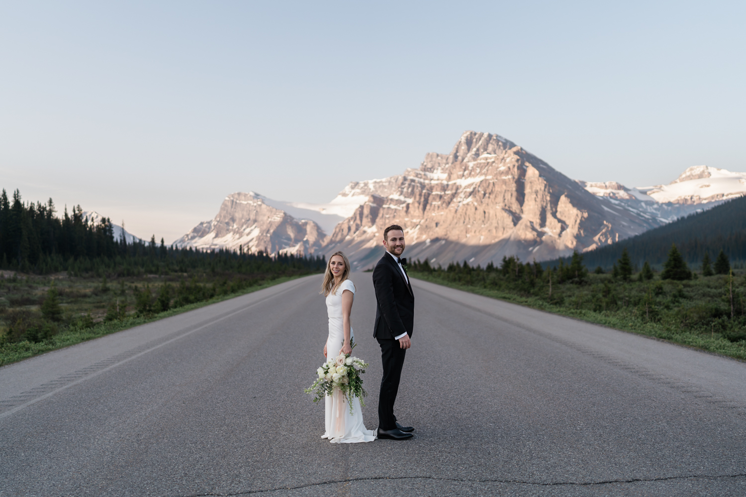 Bride and groom stand back to back during sunrise looking at the camera on the Icefields Parkway in Banff National Park