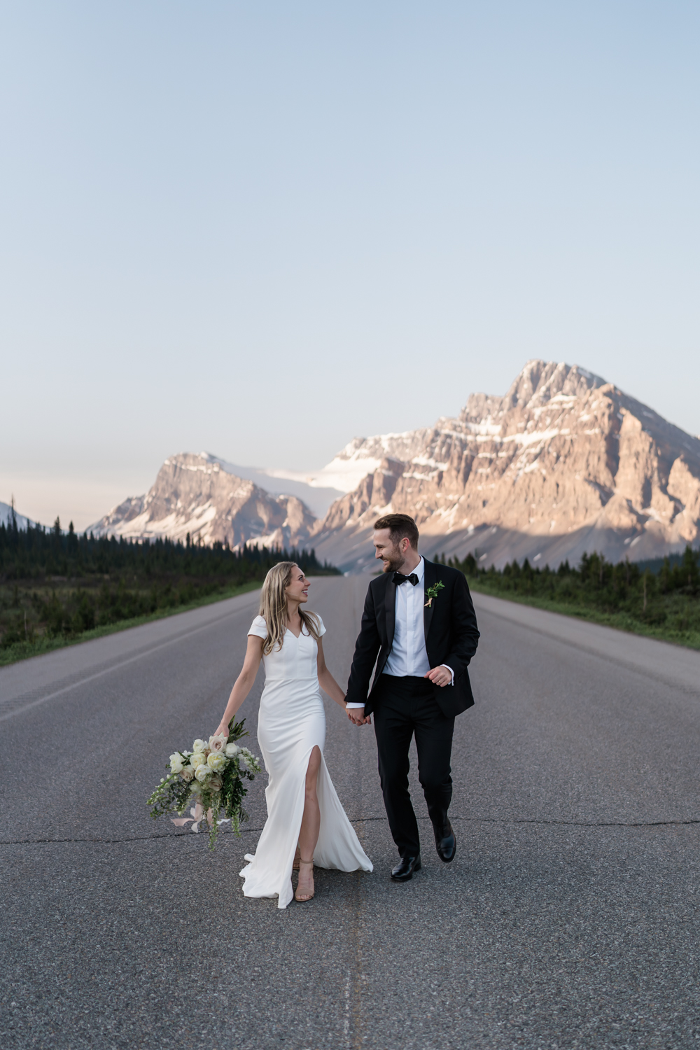 bride holding flowers and holding hands wth groom run down the road looking at each other on the Icefields Parkway in Banff National Park