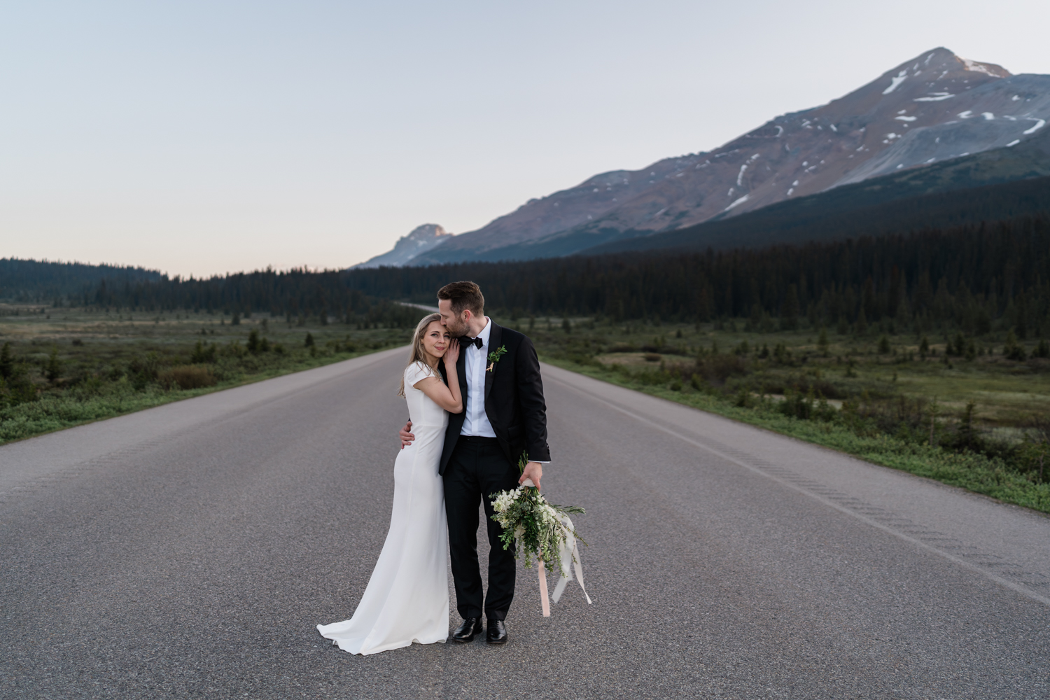 groom holds flowers and kisses head of bride while she looks at the camera and smiles on the  Icefields Parkway in Banff National Park