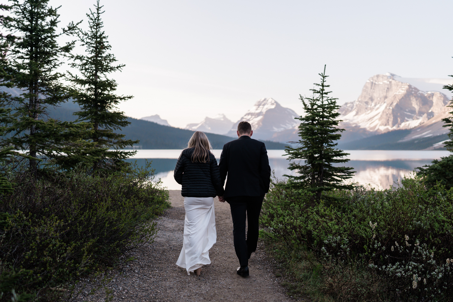 bride and groom walk through trees at Bow Lake in Banff National Park at sunrise