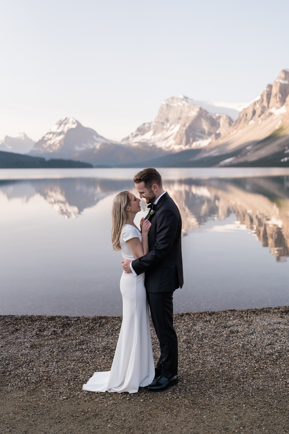 bride and groom embrace on shoreline at Bow Lake in Banff National Park at sunrise