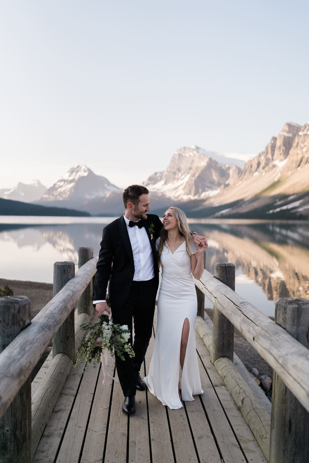 bride and groom look at each other and smile as they walk along the bridge at Bow Lake in Banff National Park at sunrise