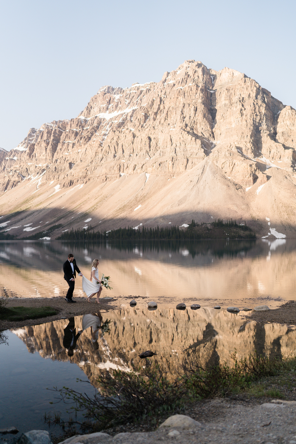 groom holds bride dress up as she walks along the shoreline at Bow Lake in Banff National Park at sunrise with the mountain reflecting in the foreground