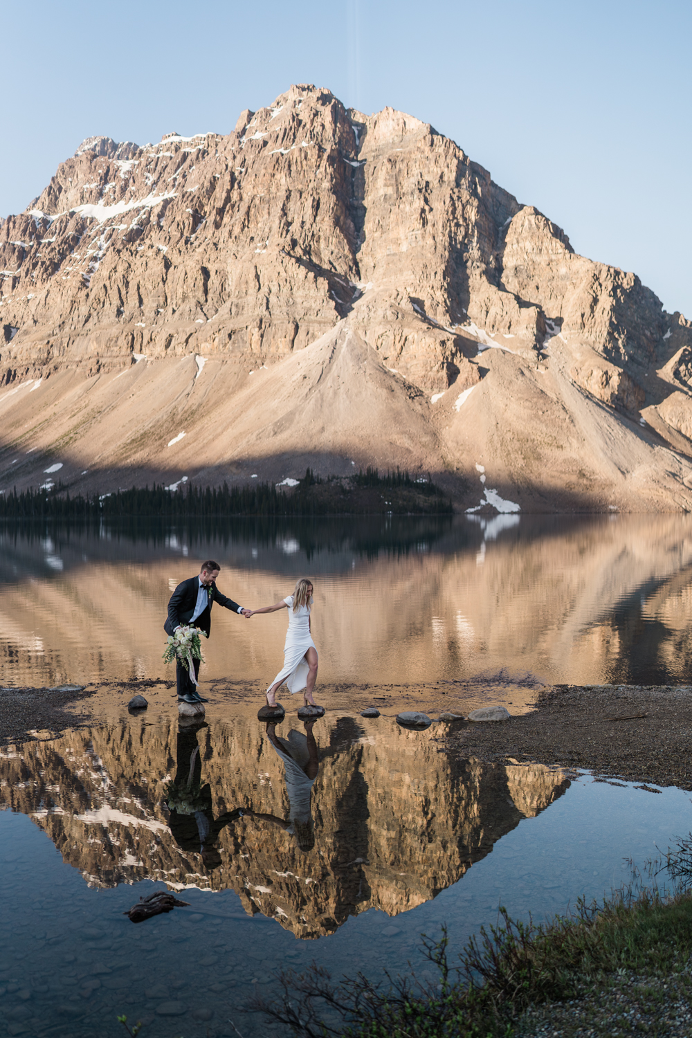 bride and groom hop over the rocks at Bow Lake in Banff National Park at sunrise with the mountain reflection
