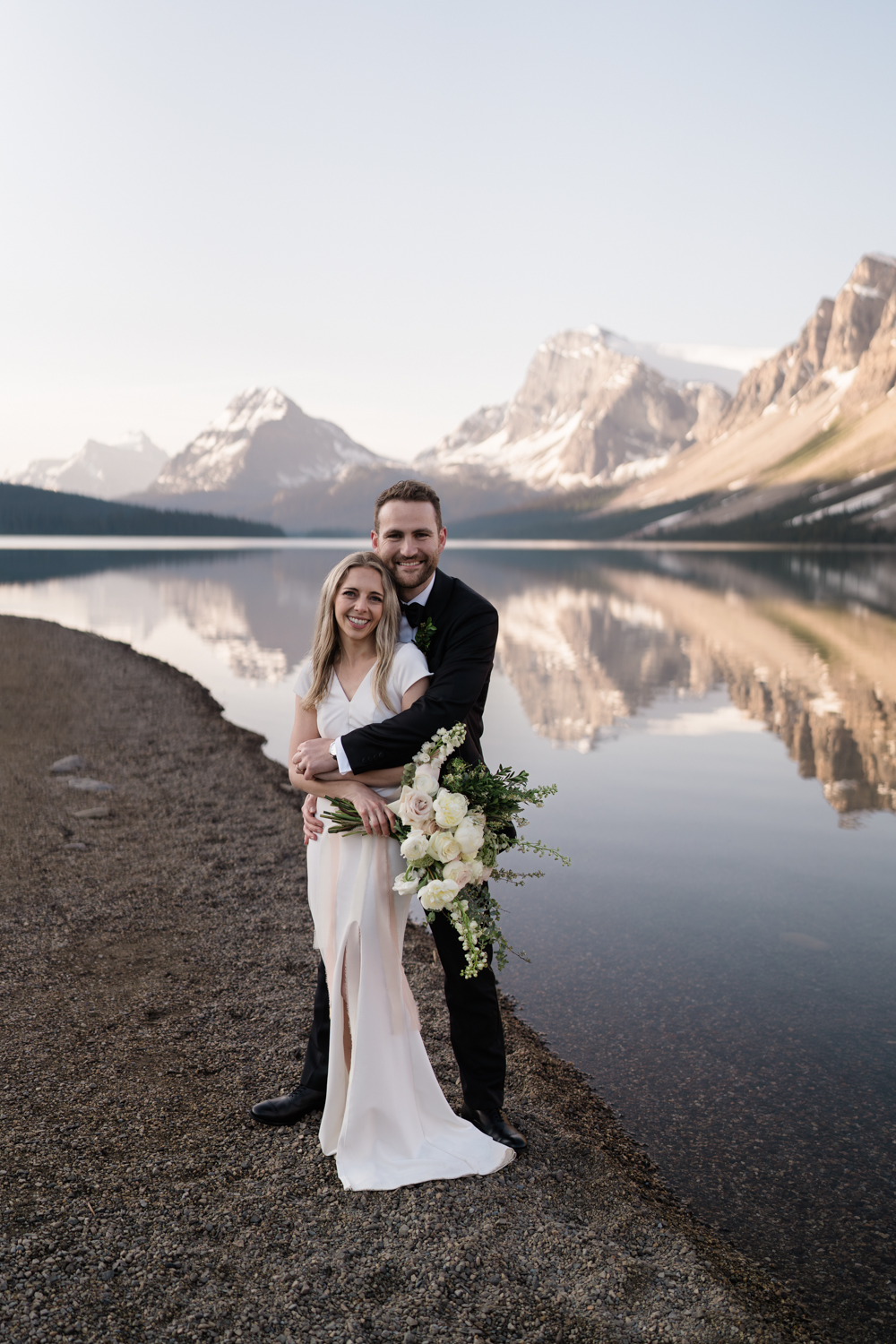 bride and groom stand and smile at the camera at Bow Lake in Banff National Park at sunrise