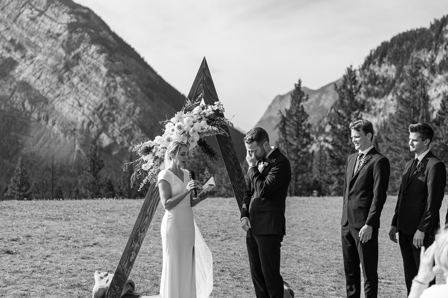 black and white photo of groom crying and wiping his eyes while bride reads vows during ceremony at Tunnel Mountain in Banff 