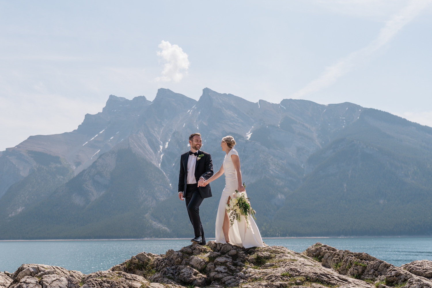 bride and groom hold hands and stand on rock at Lake Minnewanka in Banff National Park