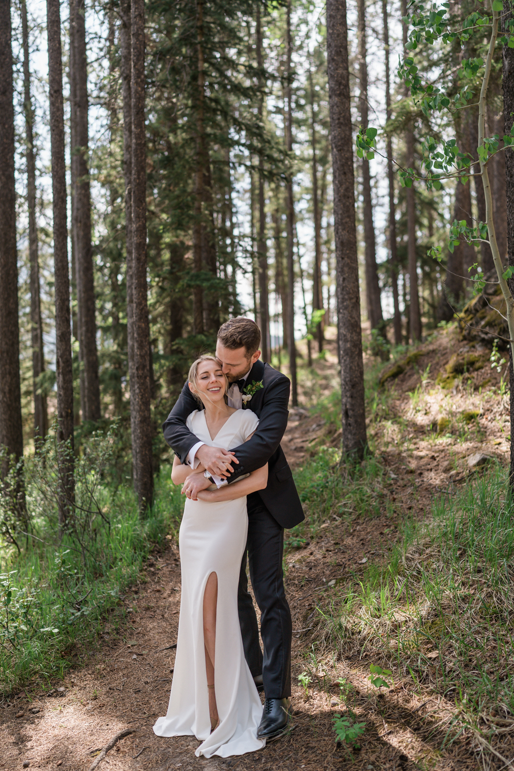 groom embraces bride from behind in the trees at Lake Minnewanka in Banff National Park