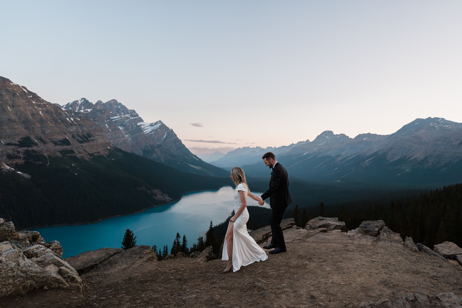 bride and groom holds hand walking at viewpoint above lake for their sunrise wedding in banff