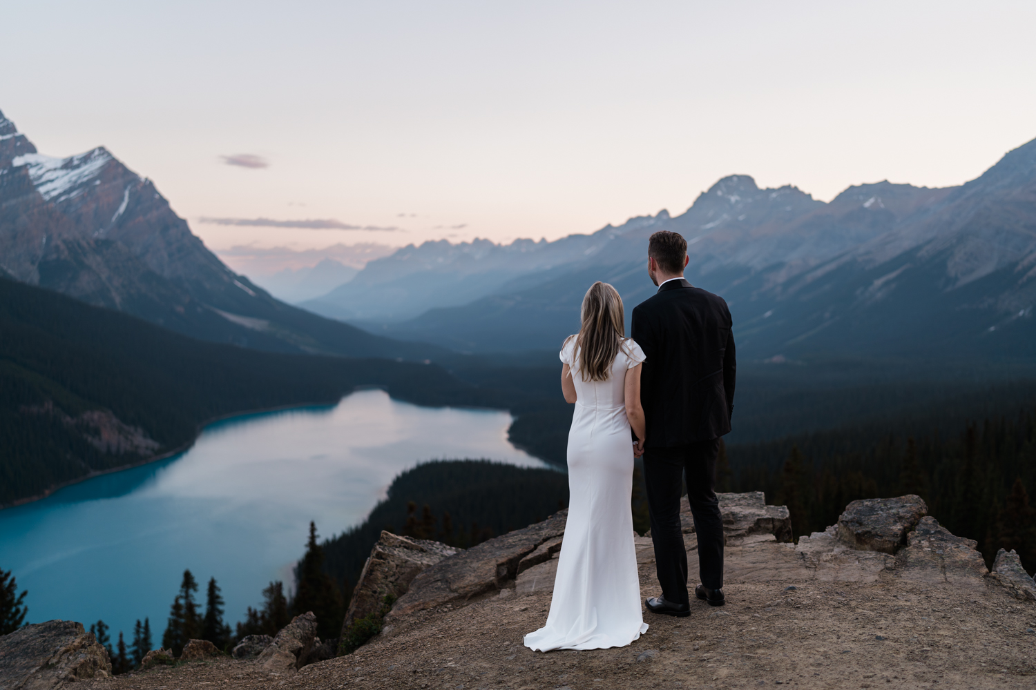 bride and groom hold hands with their backs to the camera looking out over Peyto Lake as the sun comes up behind them
