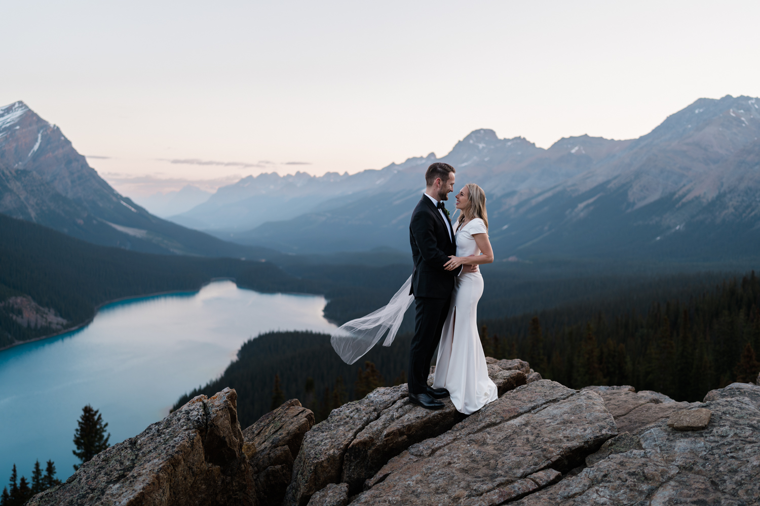 Bride and groom hold onto each other and smile as her veil blows in the wind during their Banff National Park sunrise elopement